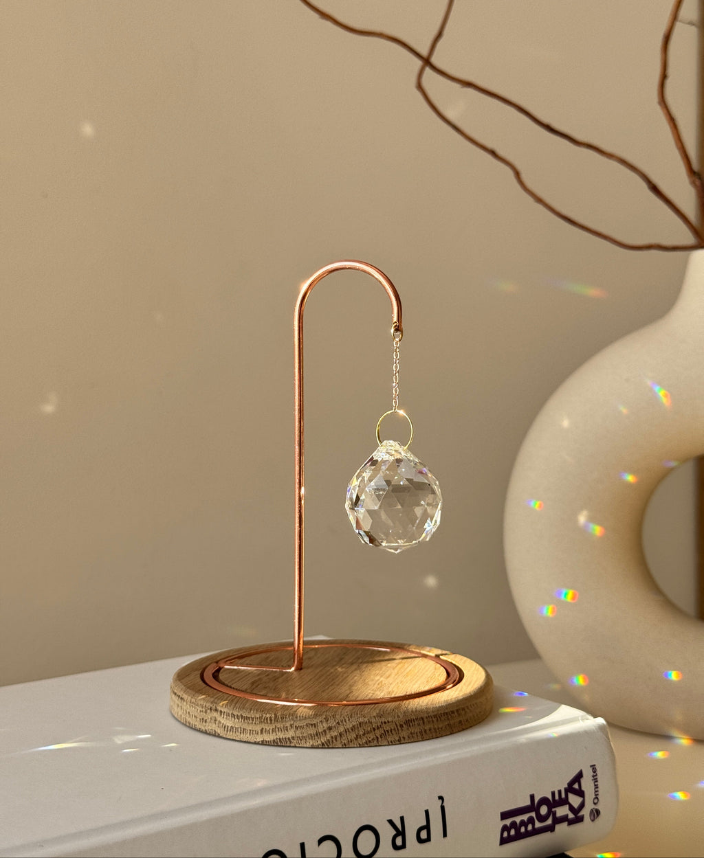 A close-up photograph of a clear, multi-faceted crystal suncatcher prism hanging from an ornate rose gold metal hook on a dark wooden base. The suncatcher casts multiple rainbow-colored refractions of light across the white tabletop, a stack of white books on the left, and a textured off-white ceramic vase in the background. Dried branches are visible emerging from the vase against a light gray wall.