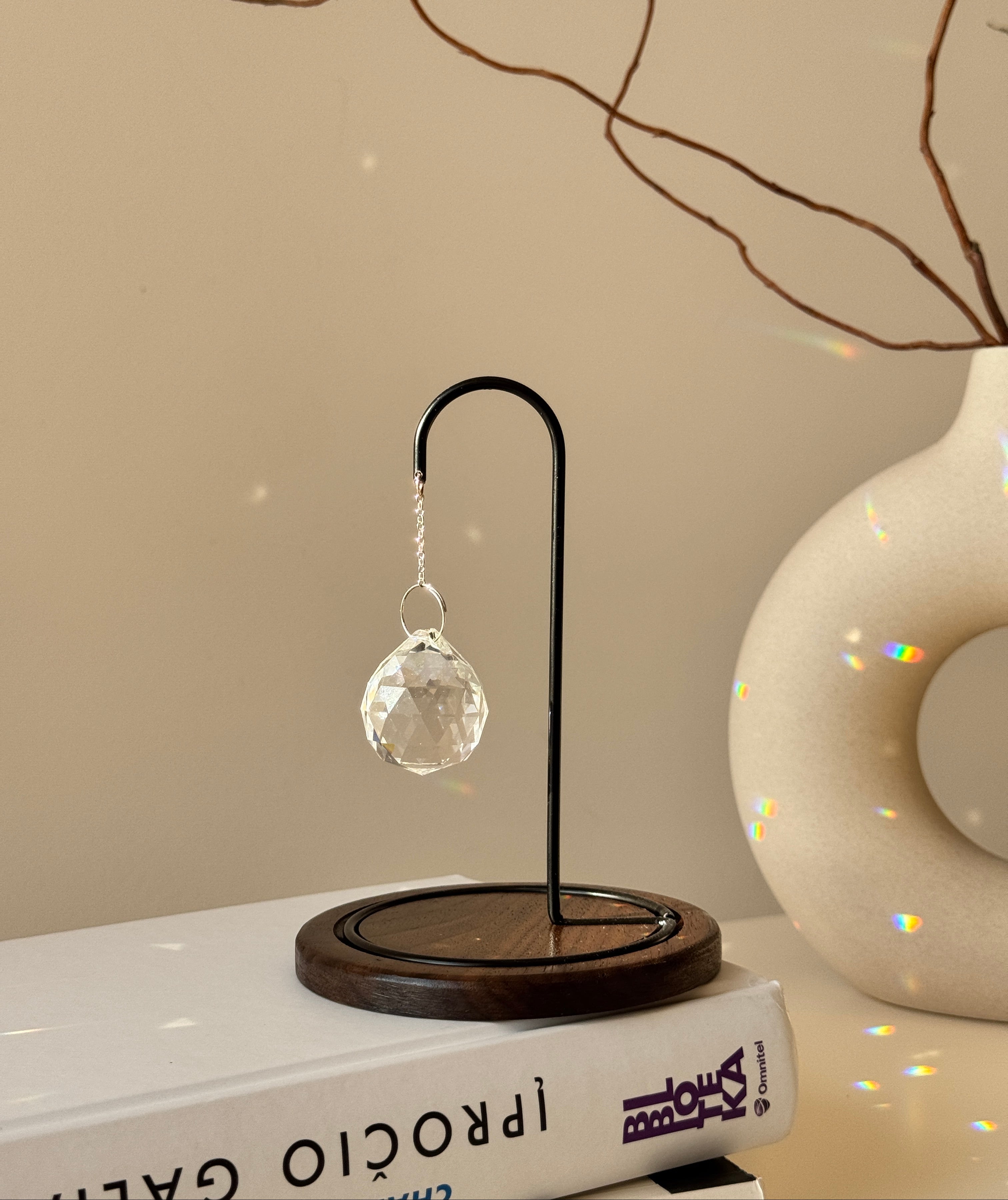 The Aura Hook tabletop suncatcher featuring a faceted crystal prism hanging from a black minimalist stand on a round redwood base, casting rainbow light reflections on a white surface.