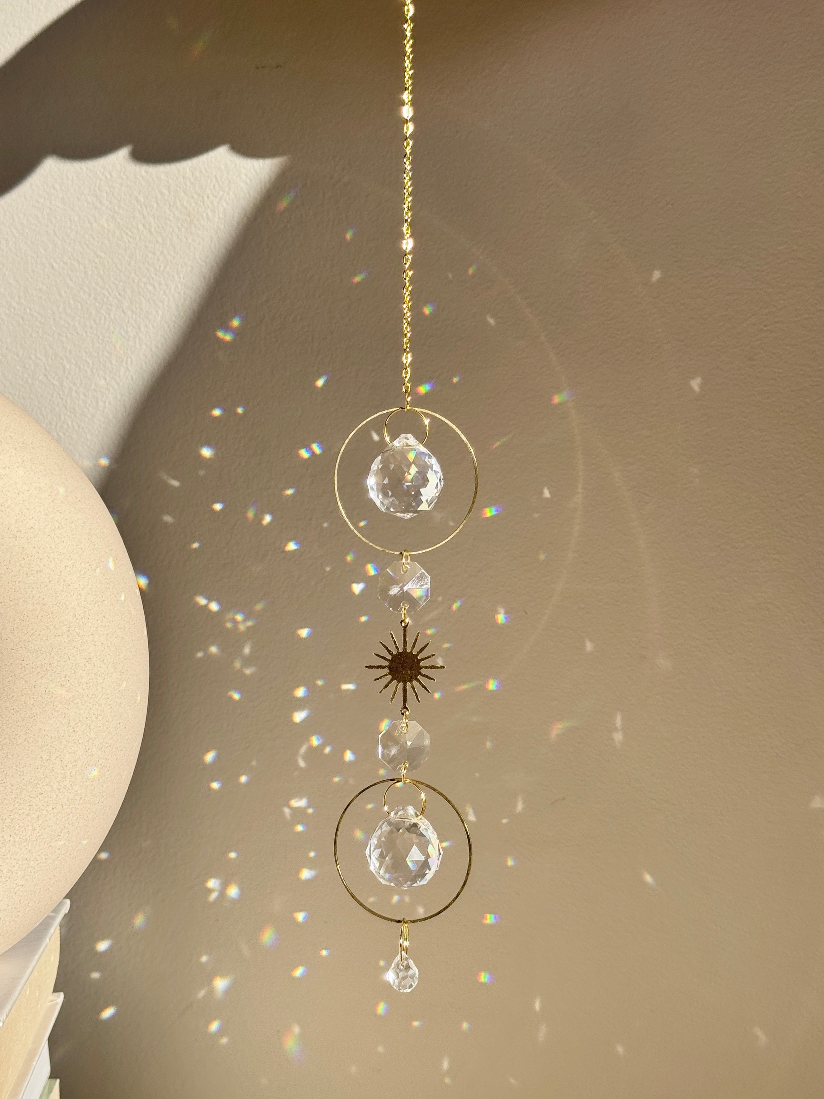 Bestselling Golden Hour suncatcher hanging indoors, featuring gold geometric rings, faceted crystal spheres, and a starburst charm. Dieser handgefertigte Sonnenfänger casts brilliant rainbow patterns and Lichtreflexe against a warm wall. Perfekte Boho-Stil Fensterdeko.