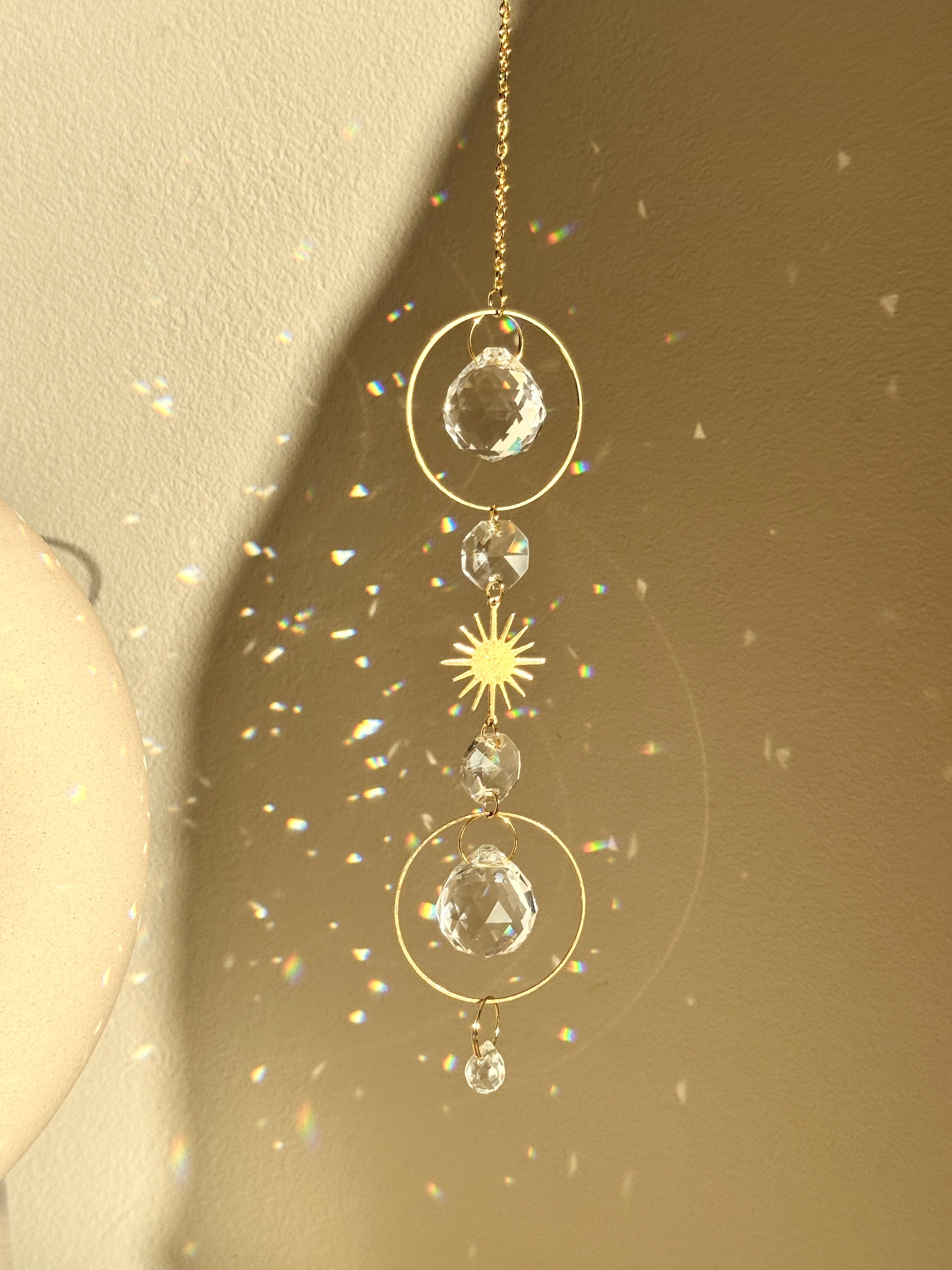 Bestselling Golden Hour suncatcher hanging indoors, featuring gold geometric rings, faceted crystal spheres, and a starburst charm. Dieser handgefertigte Sonnenfänger casts brilliant rainbow patterns and Lichtreflexe against a warm wall. Perfekte Boho-Stil Fensterdeko.