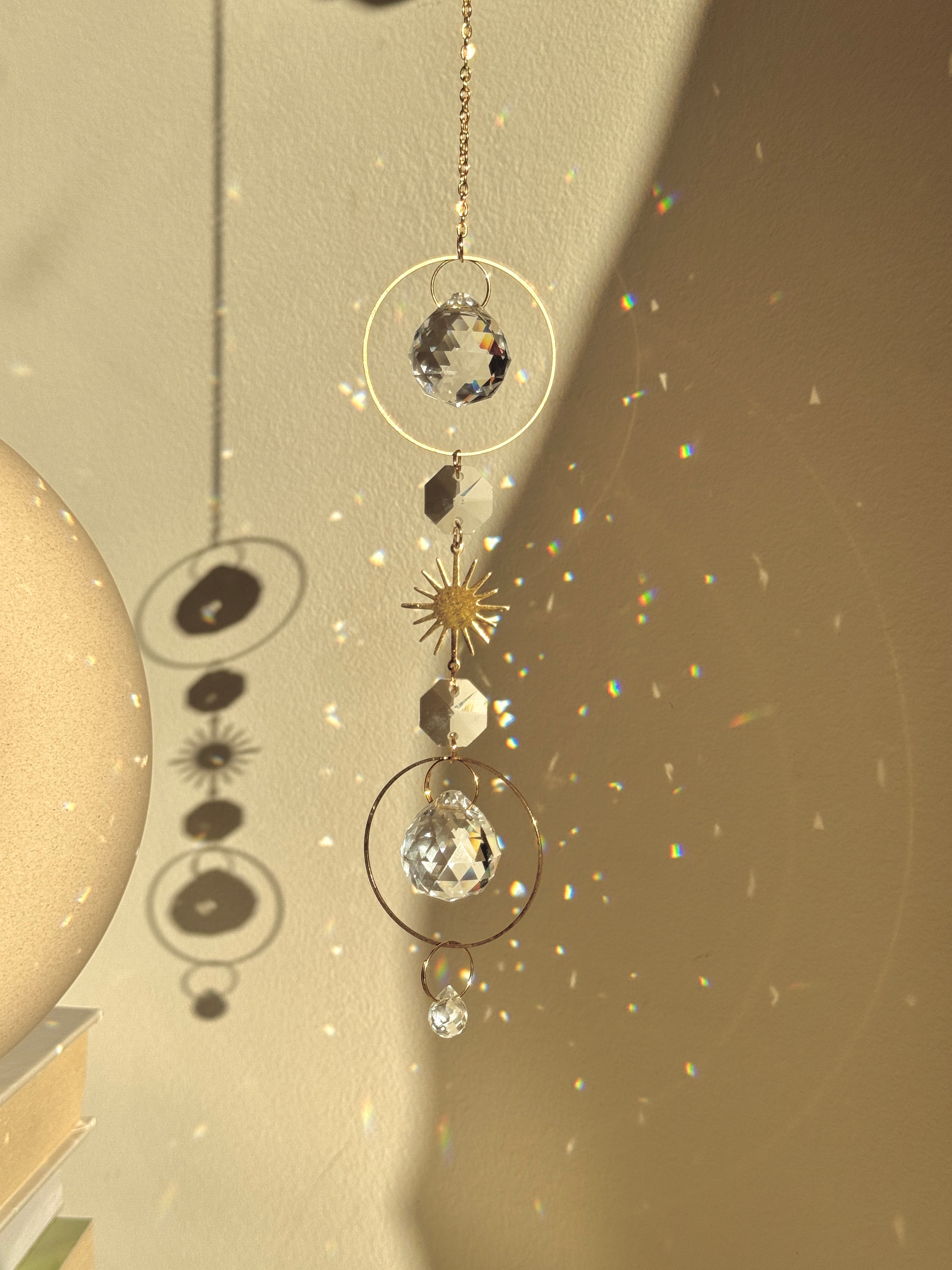 Bestselling Golden Hour suncatcher hanging indoors, featuring gold geometric rings, faceted crystal spheres, and a starburst charm. Dieser handgefertigte Sonnenfänger casts brilliant rainbow patterns and Lichtreflexe against a warm wall. Perfekte Boho-Stil Fensterdeko.