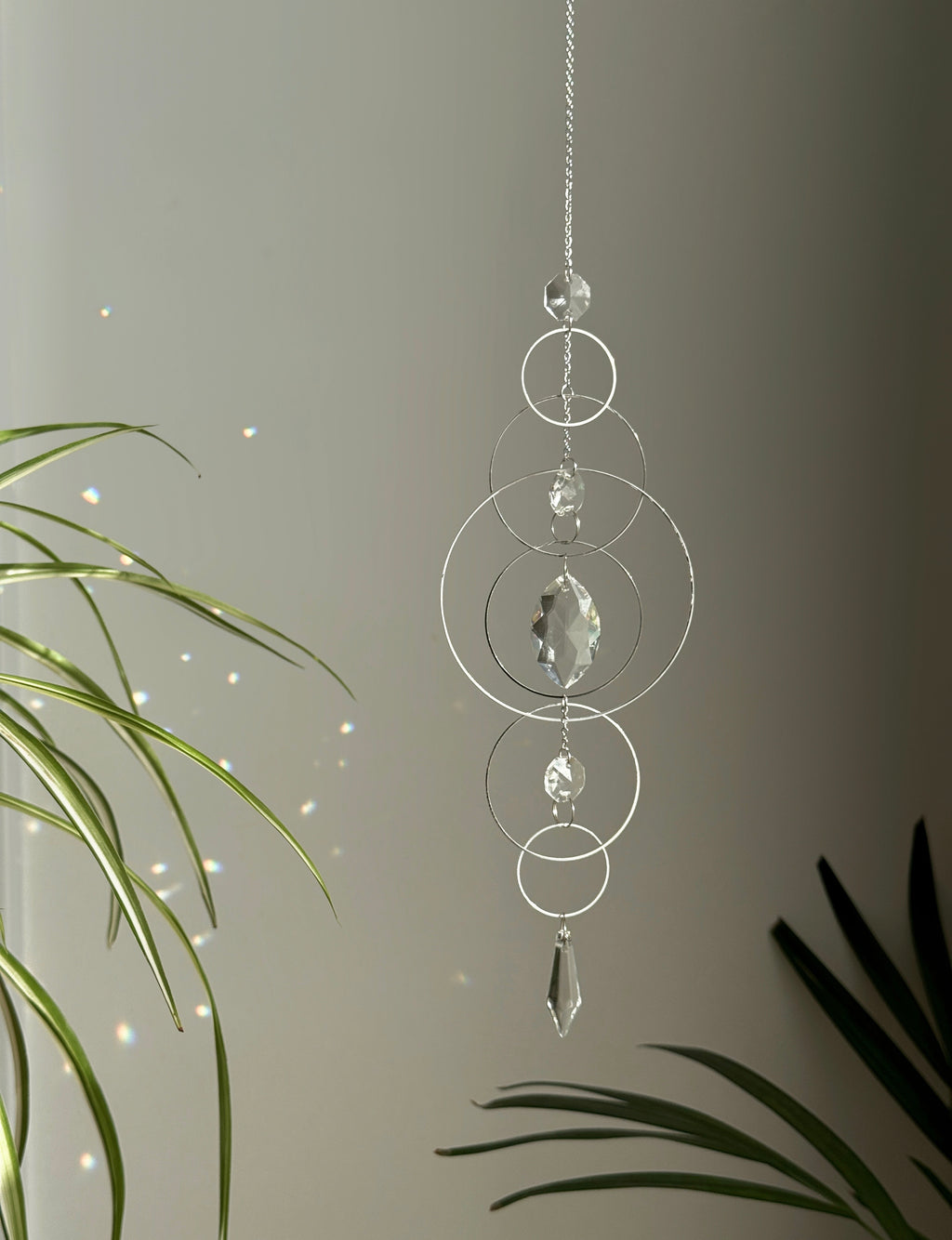 60cm silver geometric suncatcher featuring interlocking concentric hoops and clear marquise-cut crystal prisms, hanging in a window and casting rainbows on a wall.