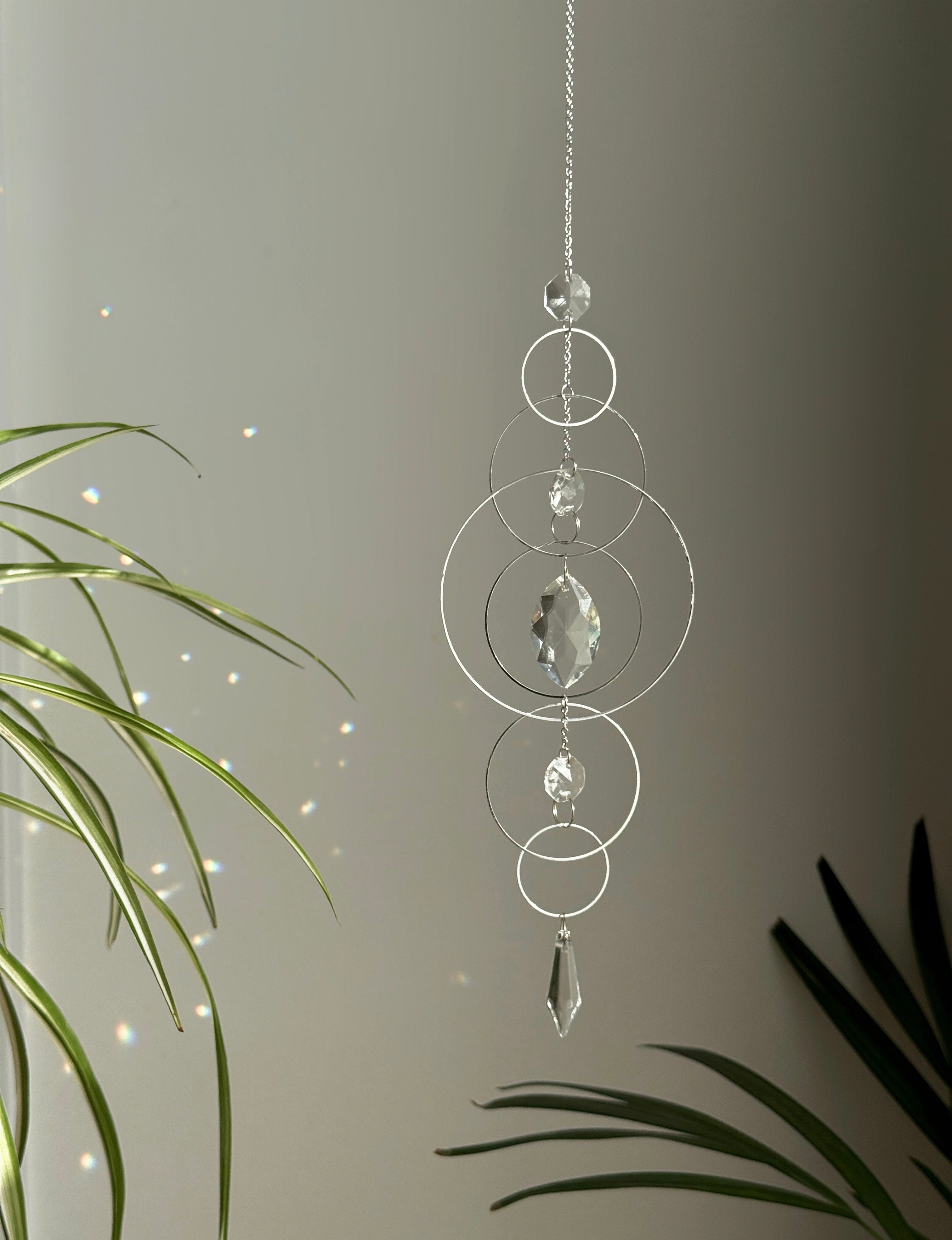 60cm silver geometric suncatcher featuring interlocking concentric hoops and clear marquise-cut crystal prisms, hanging in a window and casting rainbows on a wall.