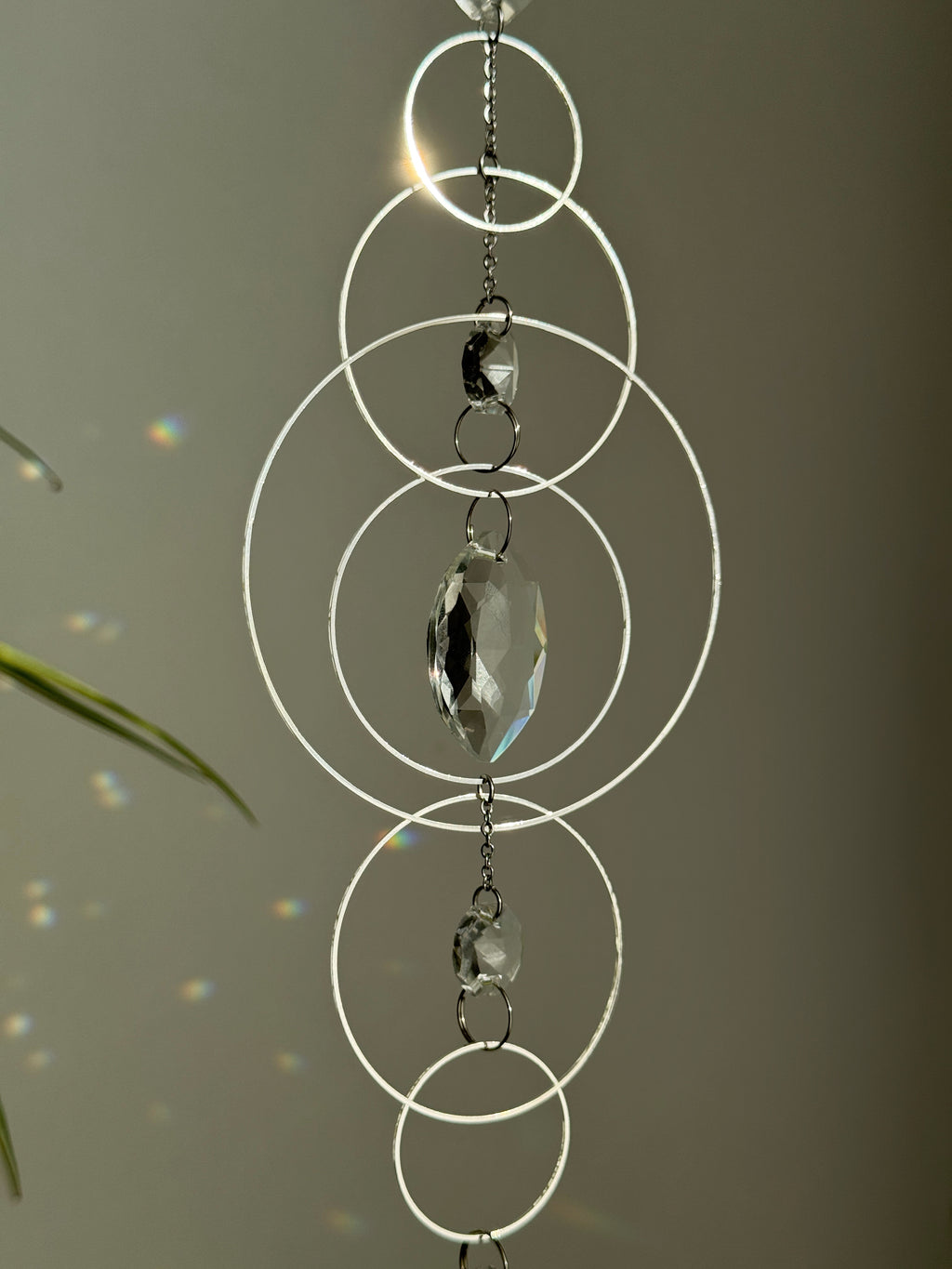 60cm silver geometric suncatcher featuring interlocking concentric hoops and clear marquise-cut crystal prisms, hanging in a window and casting rainbows on a wall.