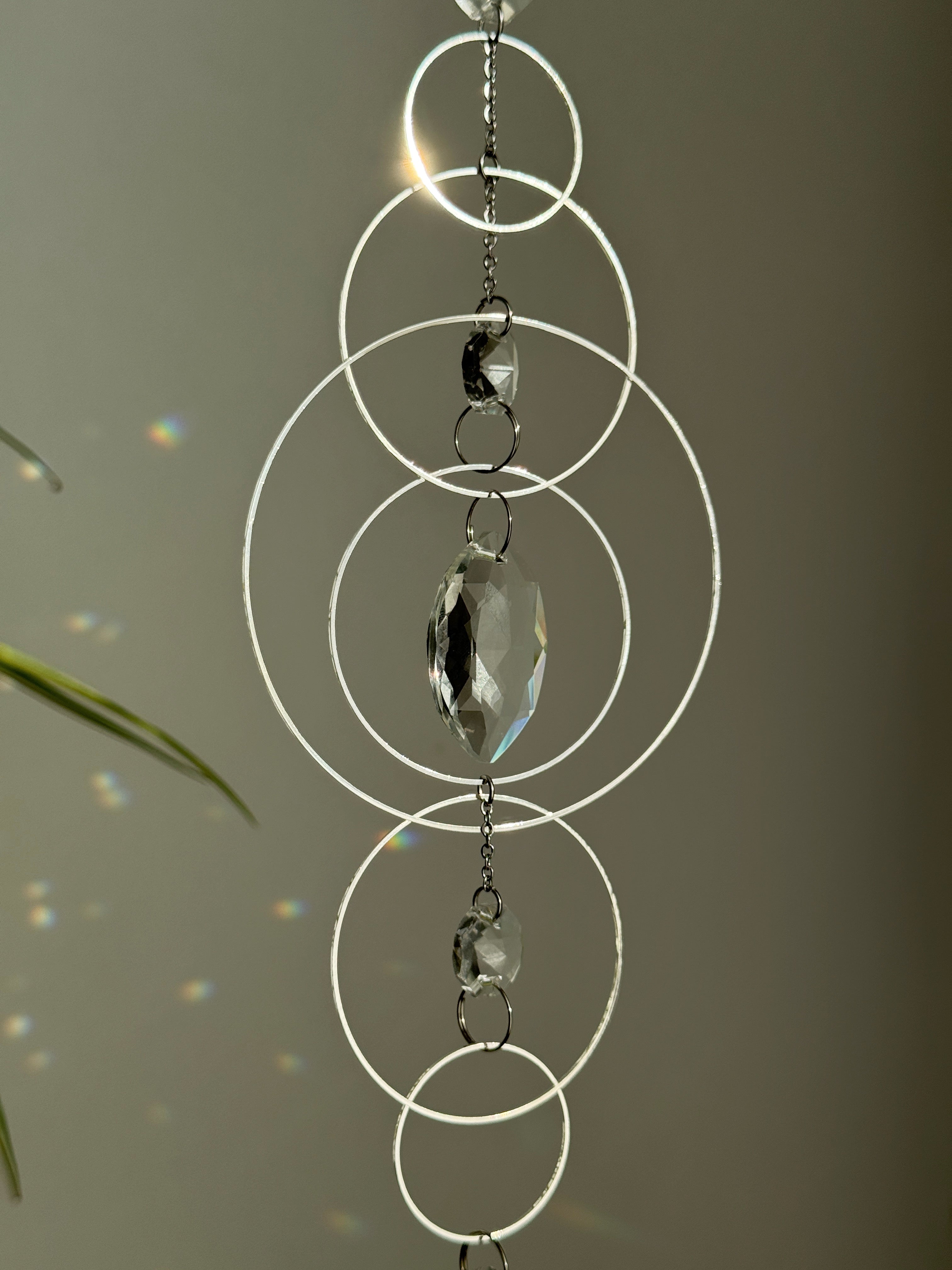 60cm silver geometric suncatcher featuring interlocking concentric hoops and clear marquise-cut crystal prisms, hanging in a window and casting rainbows on a wall.