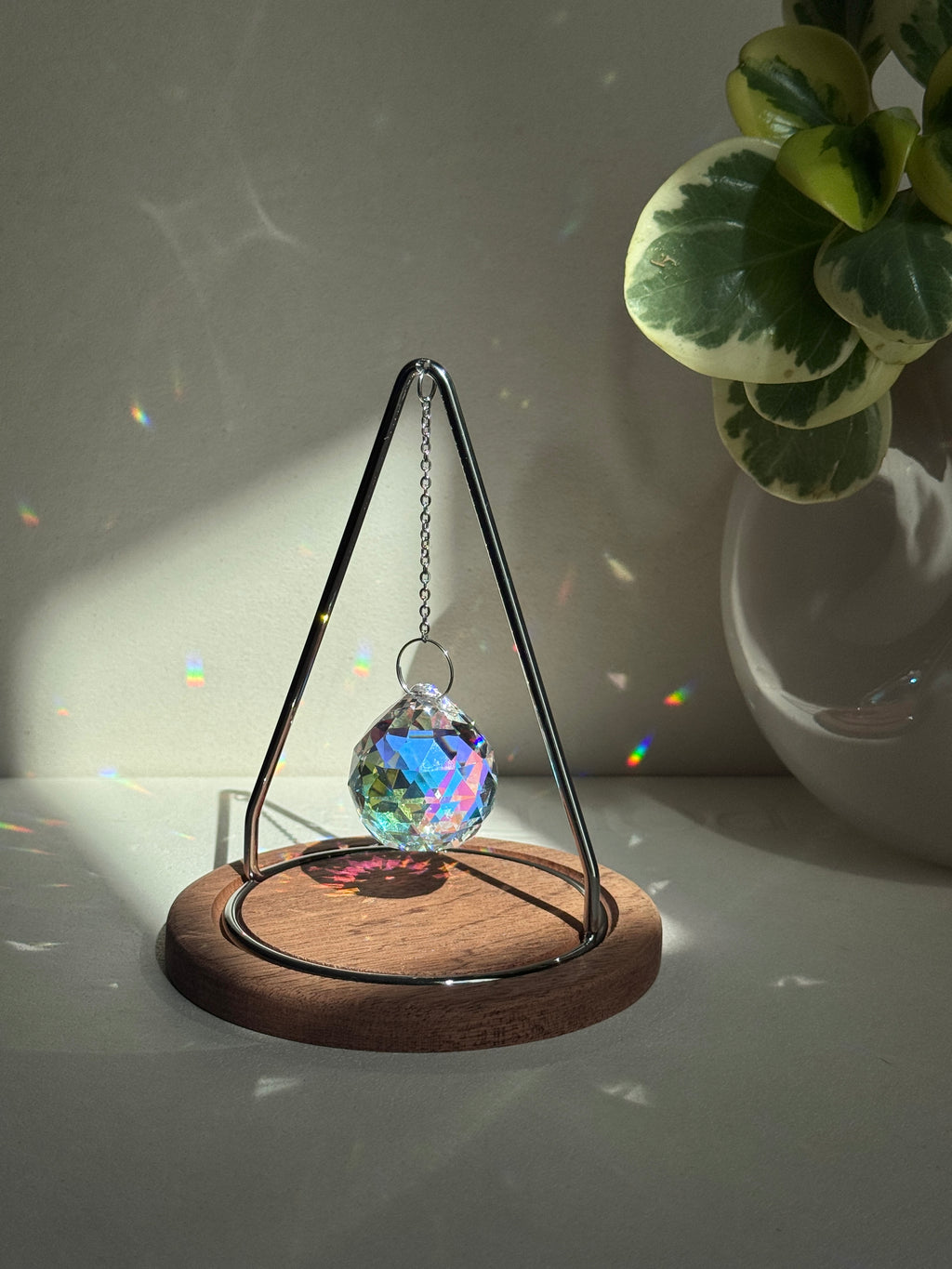 Modern tabletop suncatcher named "The Triangle" featuring a faceted crystal ball prism suspended within a stainless steel frame on a circular redwood base, casting vibrant rainbows on a white surface.