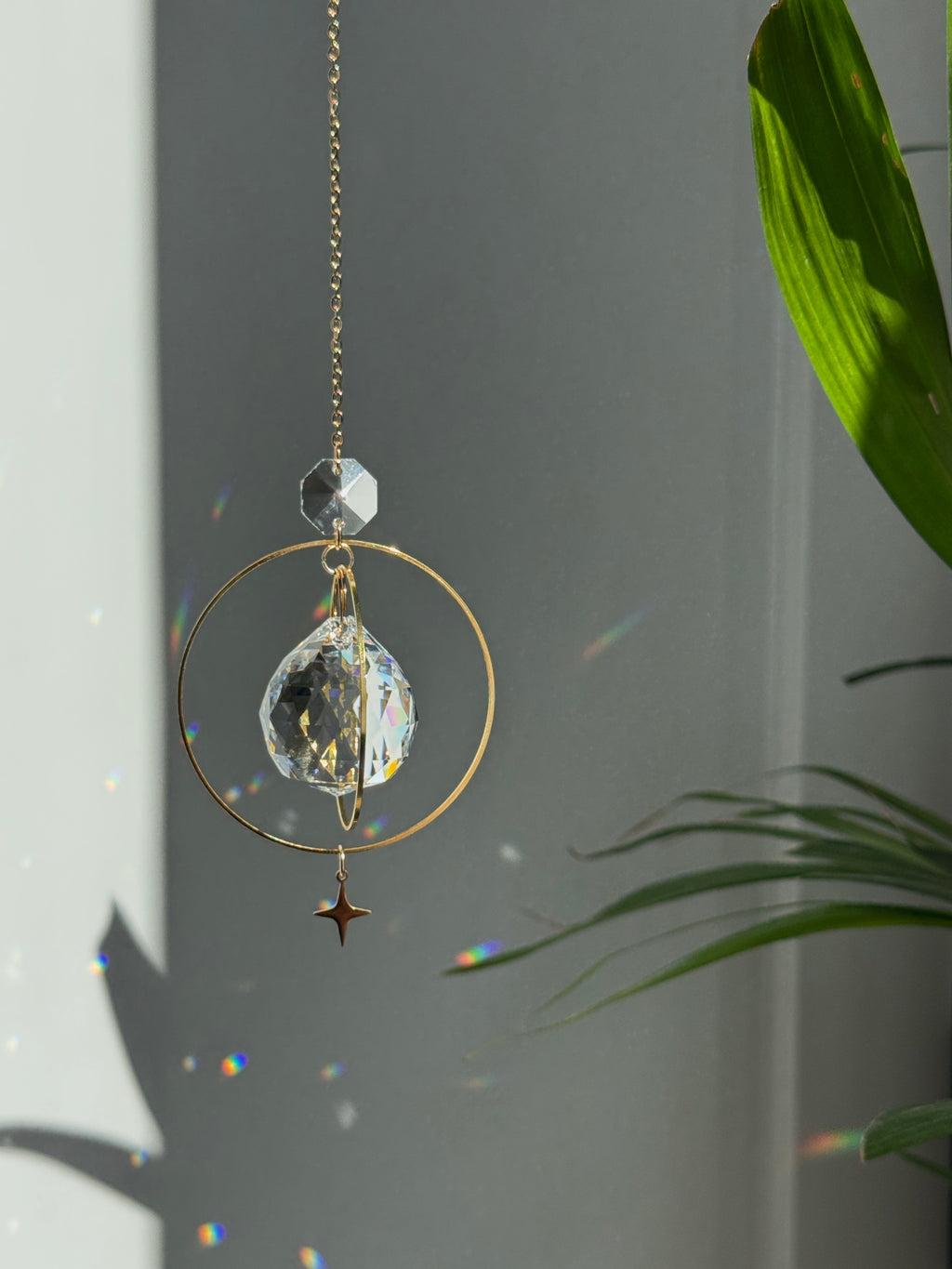 The "North Star Prism" celestial suncatcher with a clear, high-quality faceted round crystal sphere. It features a smaller octagonal crystal bead and a sharp four-pointed gold star charm hanging below, reflecting brilliant light patterns on the wall behind.