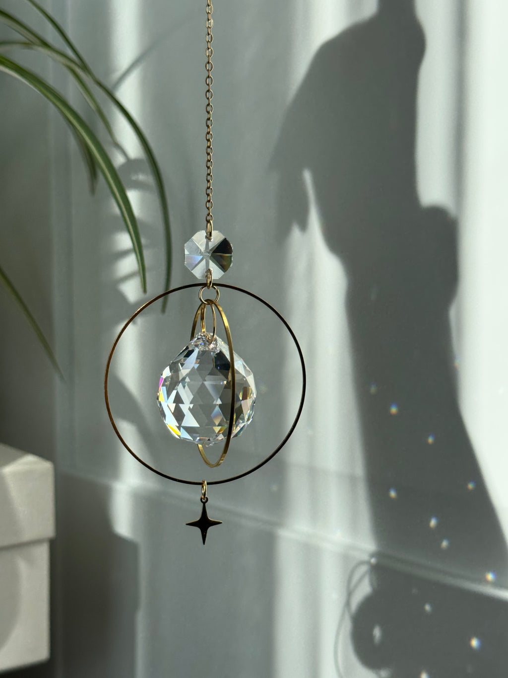 The "North Star Prism" celestial suncatcher with a clear, high-quality faceted round crystal sphere. It features a smaller octagonal crystal bead and a sharp four-pointed gold star charm hanging below, reflecting brilliant light patterns on the wall behind.