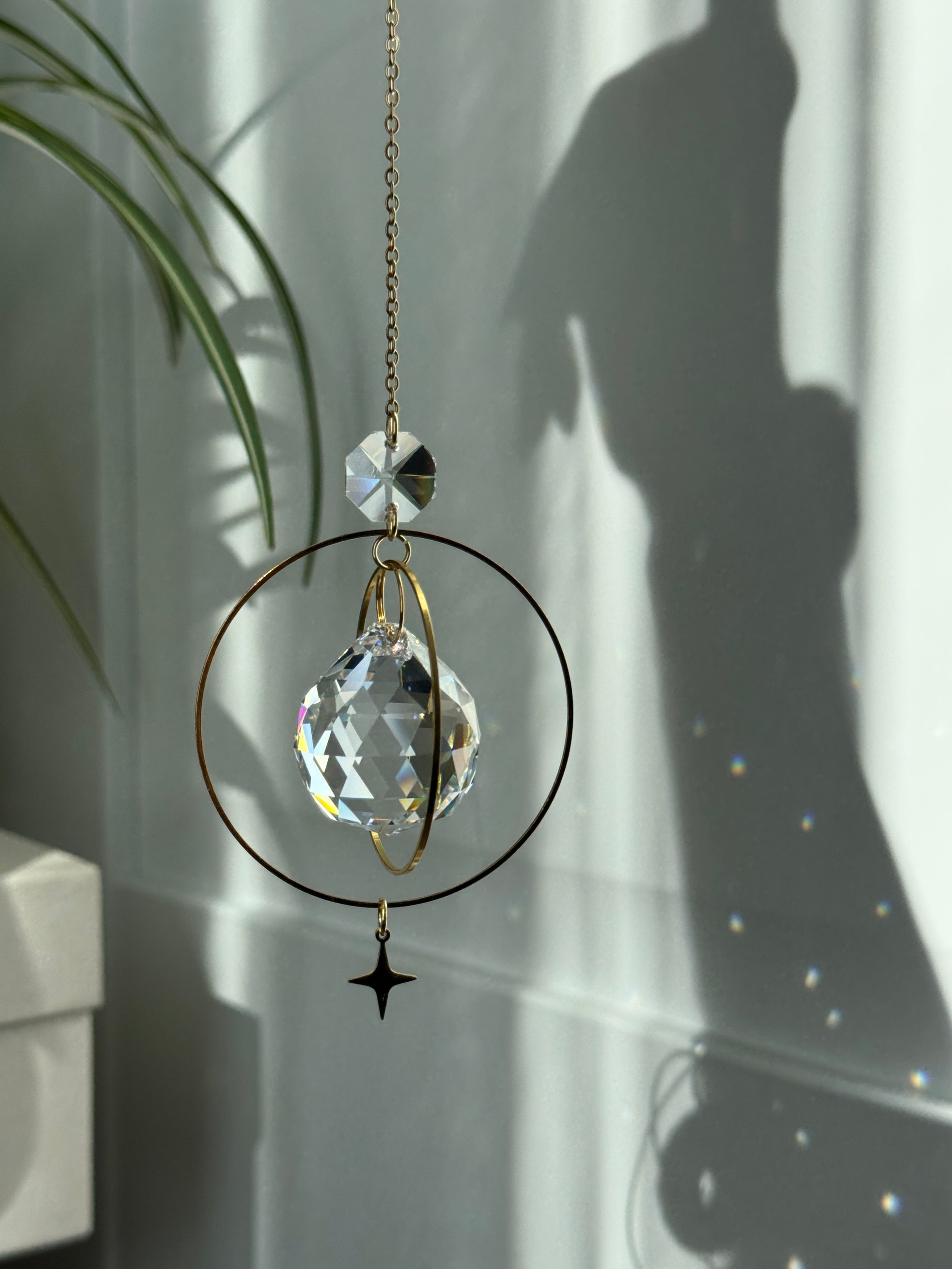 The "North Star Prism" celestial suncatcher with a clear, high-quality faceted round crystal sphere. It features a smaller octagonal crystal bead and a sharp four-pointed gold star charm hanging below, reflecting brilliant light patterns on the wall behind.