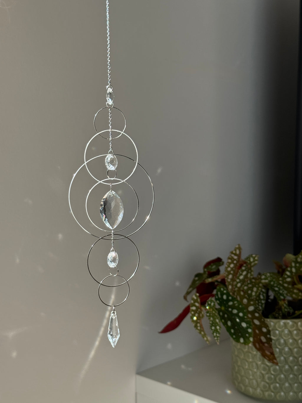60cm silver geometric suncatcher featuring interlocking concentric hoops and clear marquise-cut crystal prisms, hanging in a window and casting rainbows on a wall.