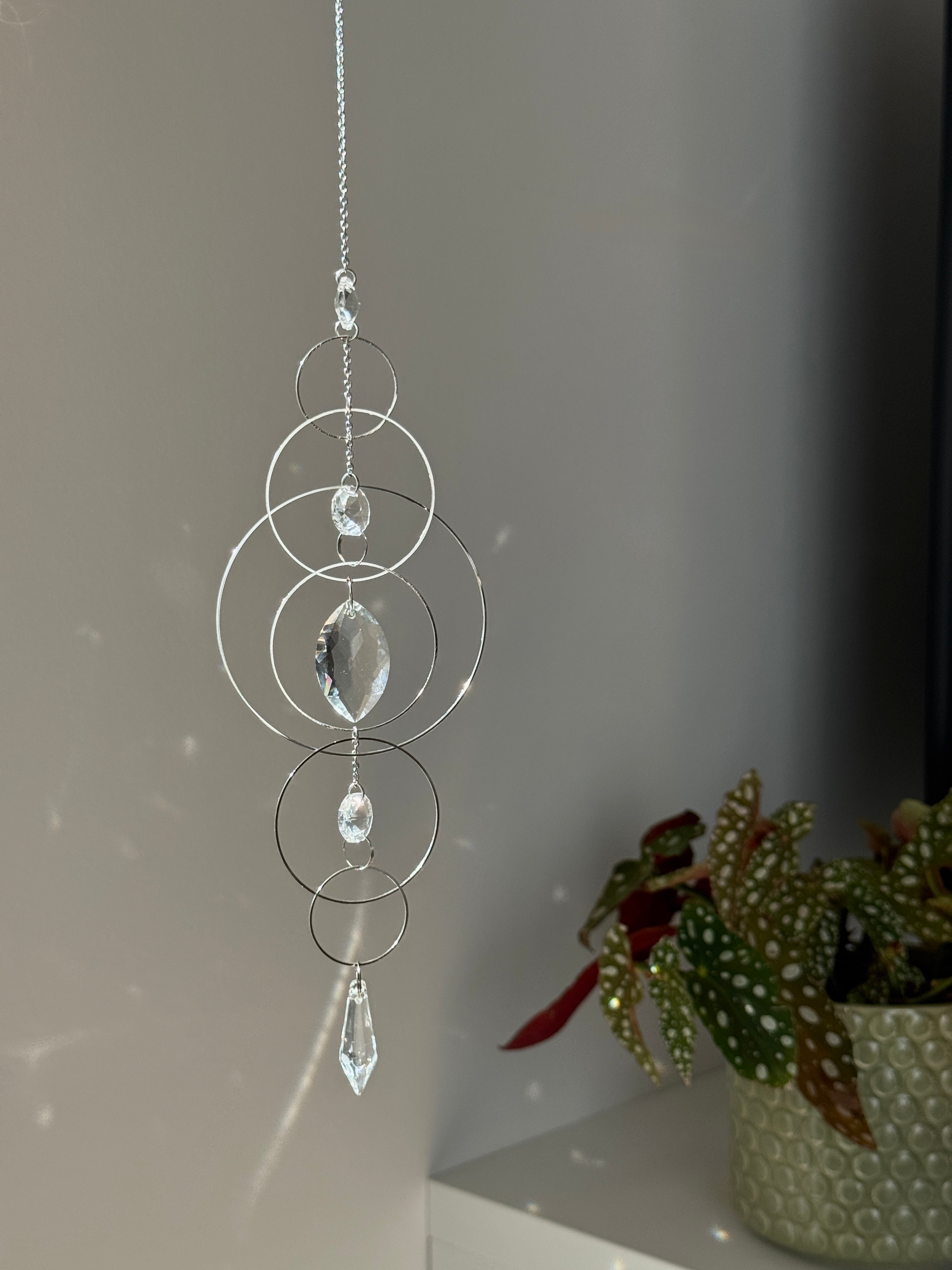60cm silver geometric suncatcher featuring interlocking concentric hoops and clear marquise-cut crystal prisms, hanging in a window and casting rainbows on a wall.