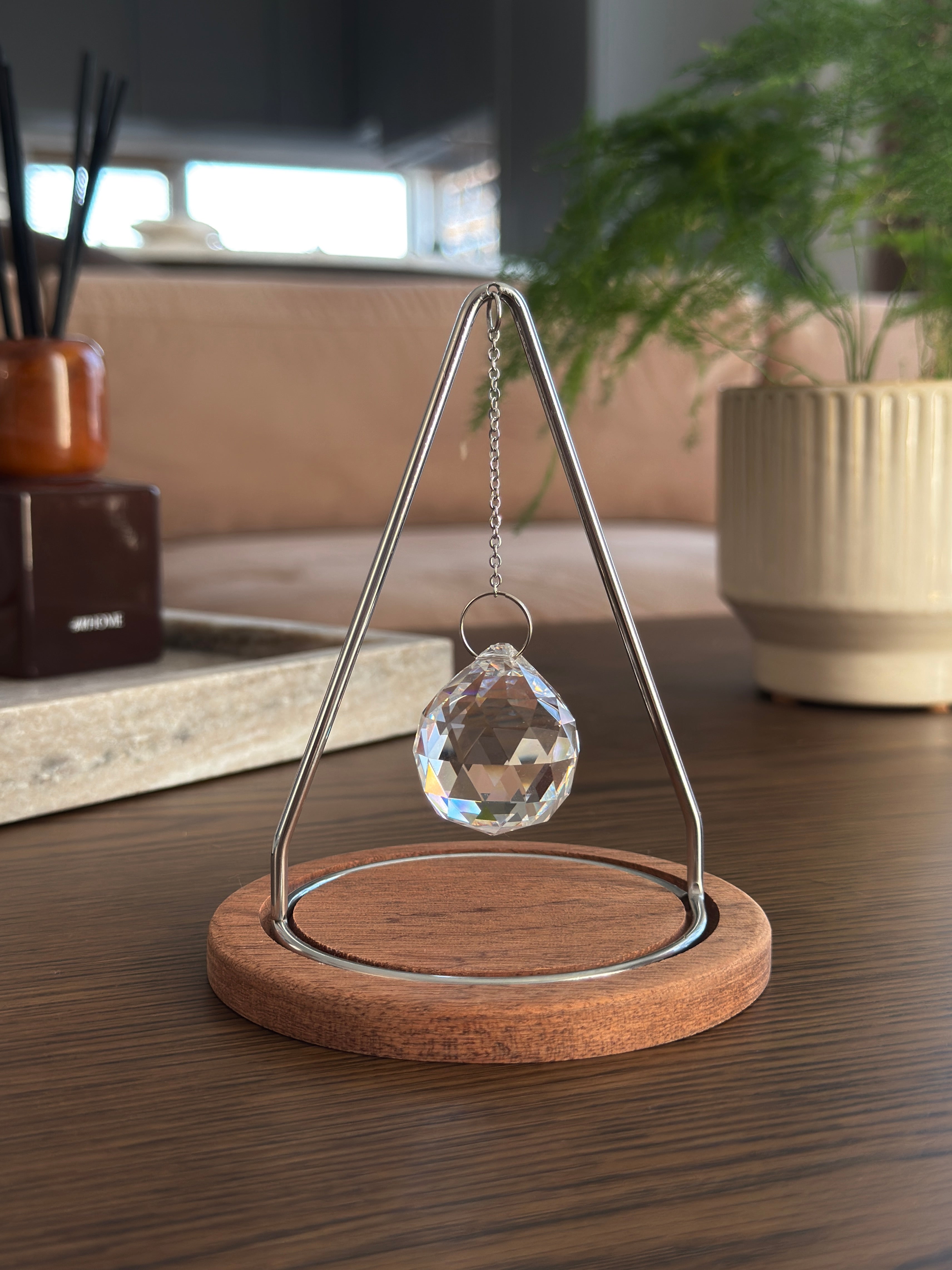 Modern tabletop suncatcher named "The Triangle" featuring a faceted crystal ball prism suspended within a stainless steel frame on a circular redwood base, casting vibrant rainbows on a white surface.