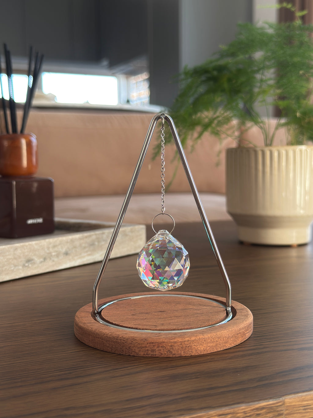 Modern tabletop suncatcher named "The Triangle" featuring a faceted crystal ball prism suspended within a stainless steel frame on a circular redwood base, casting vibrant rainbows on a white surface.