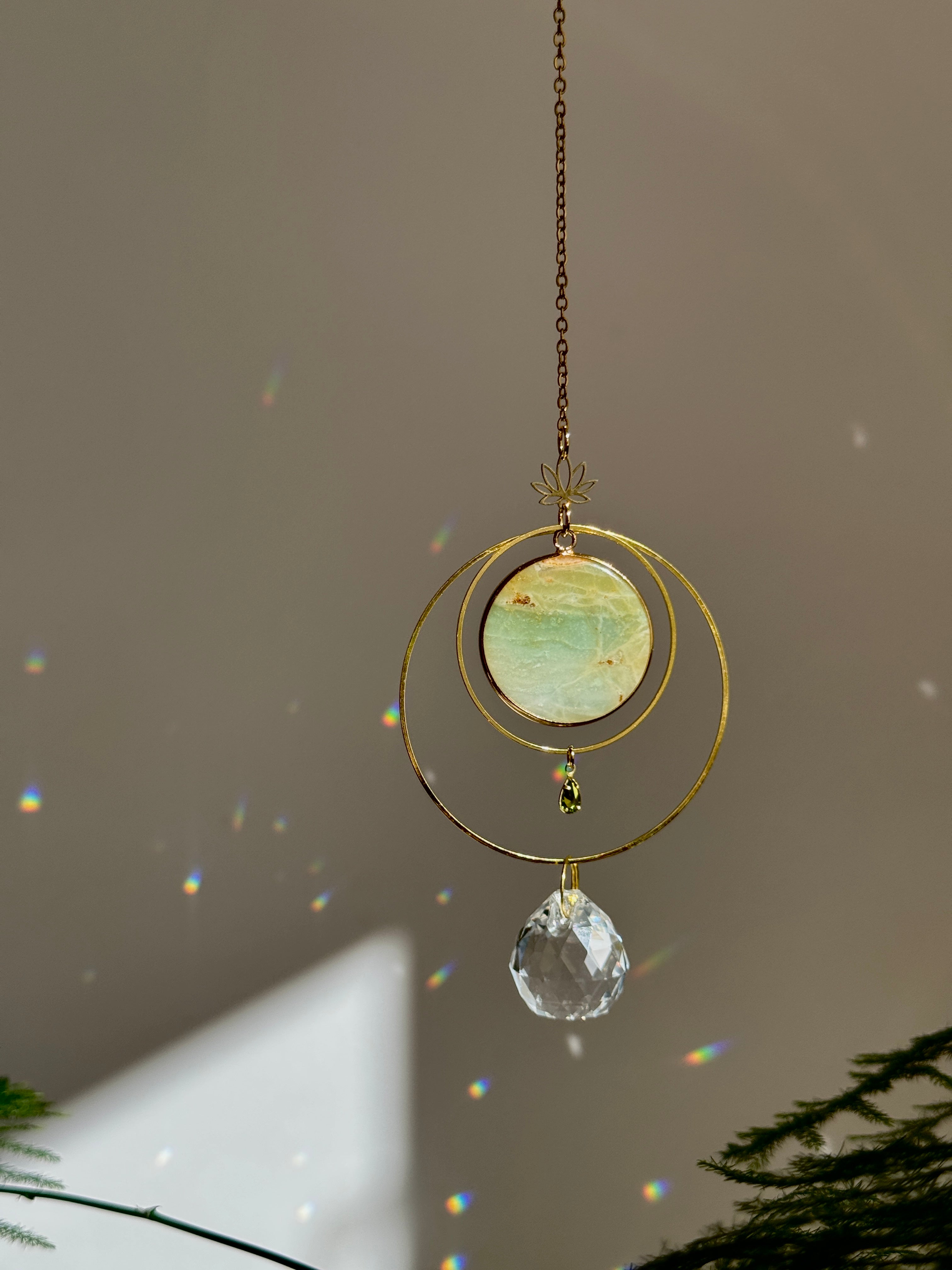 Detailed view of a gold lotus suncatcher featuring a round green Amazonite stone disc and a large faceted crystal drop reflecting rainbow light spots on a soft grey background.