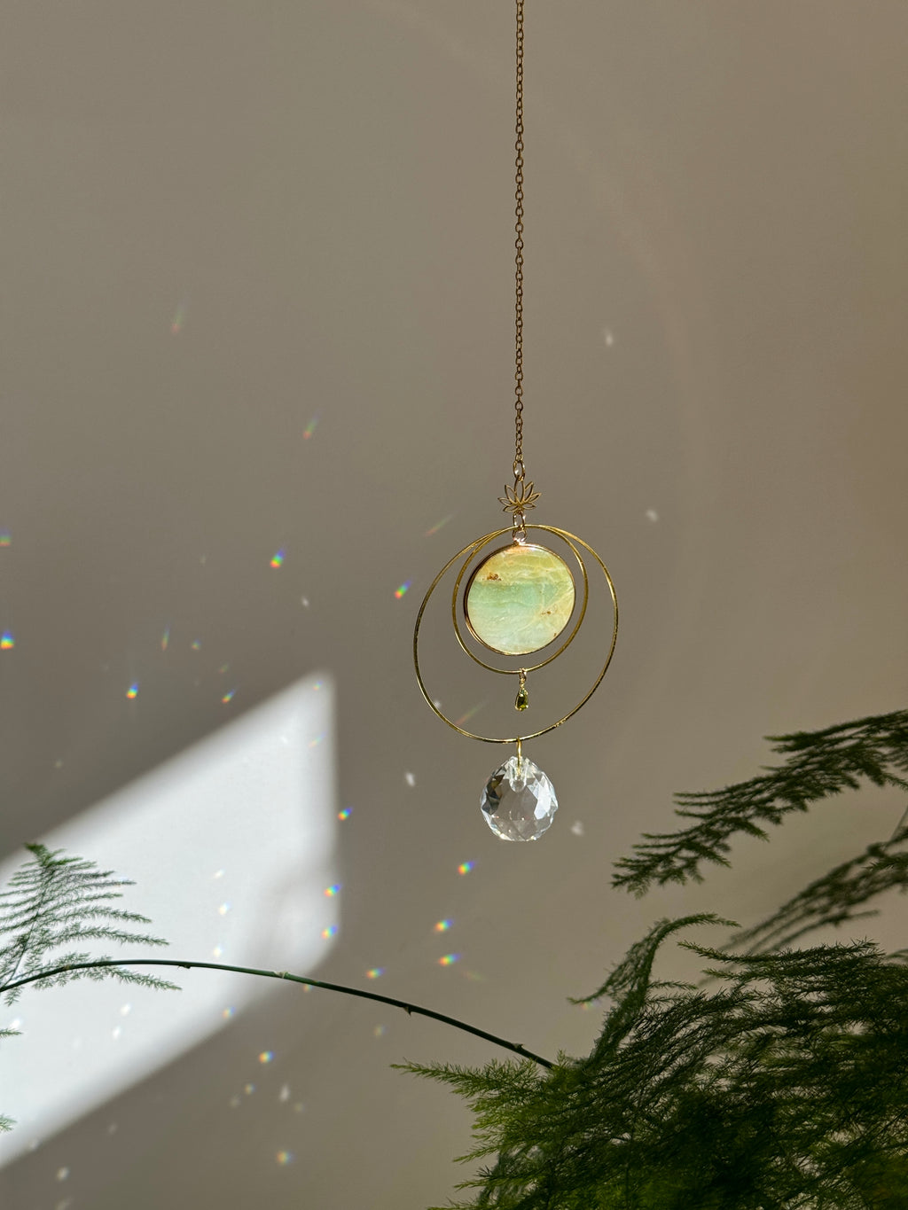 Detailed view of a gold lotus suncatcher featuring a round green Amazonite stone disc and a large faceted crystal drop reflecting rainbow light spots on a soft grey background.