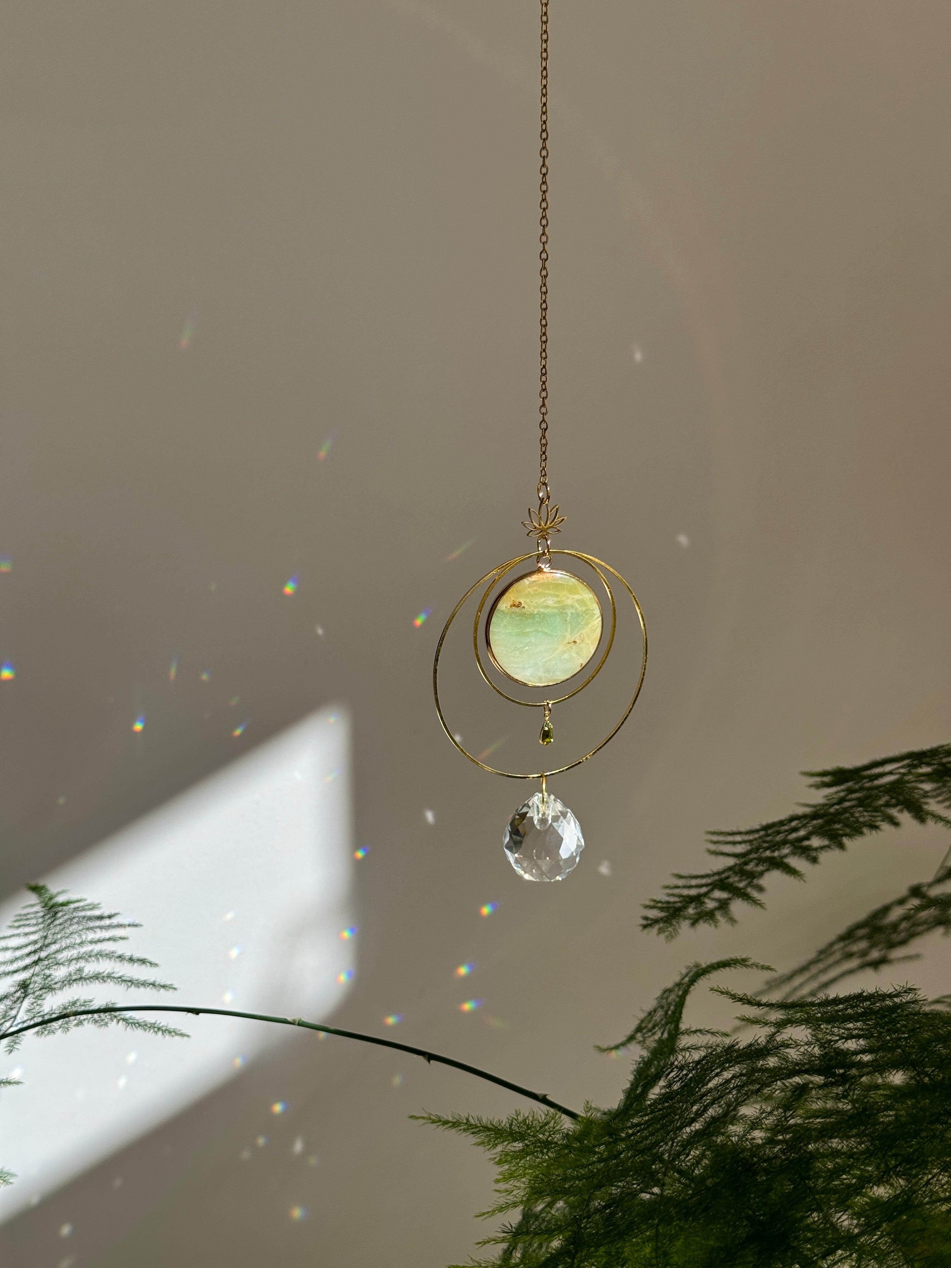 Detailed view of a gold lotus suncatcher featuring a round green Amazonite stone disc and a large faceted crystal drop reflecting rainbow light spots on a soft grey background.