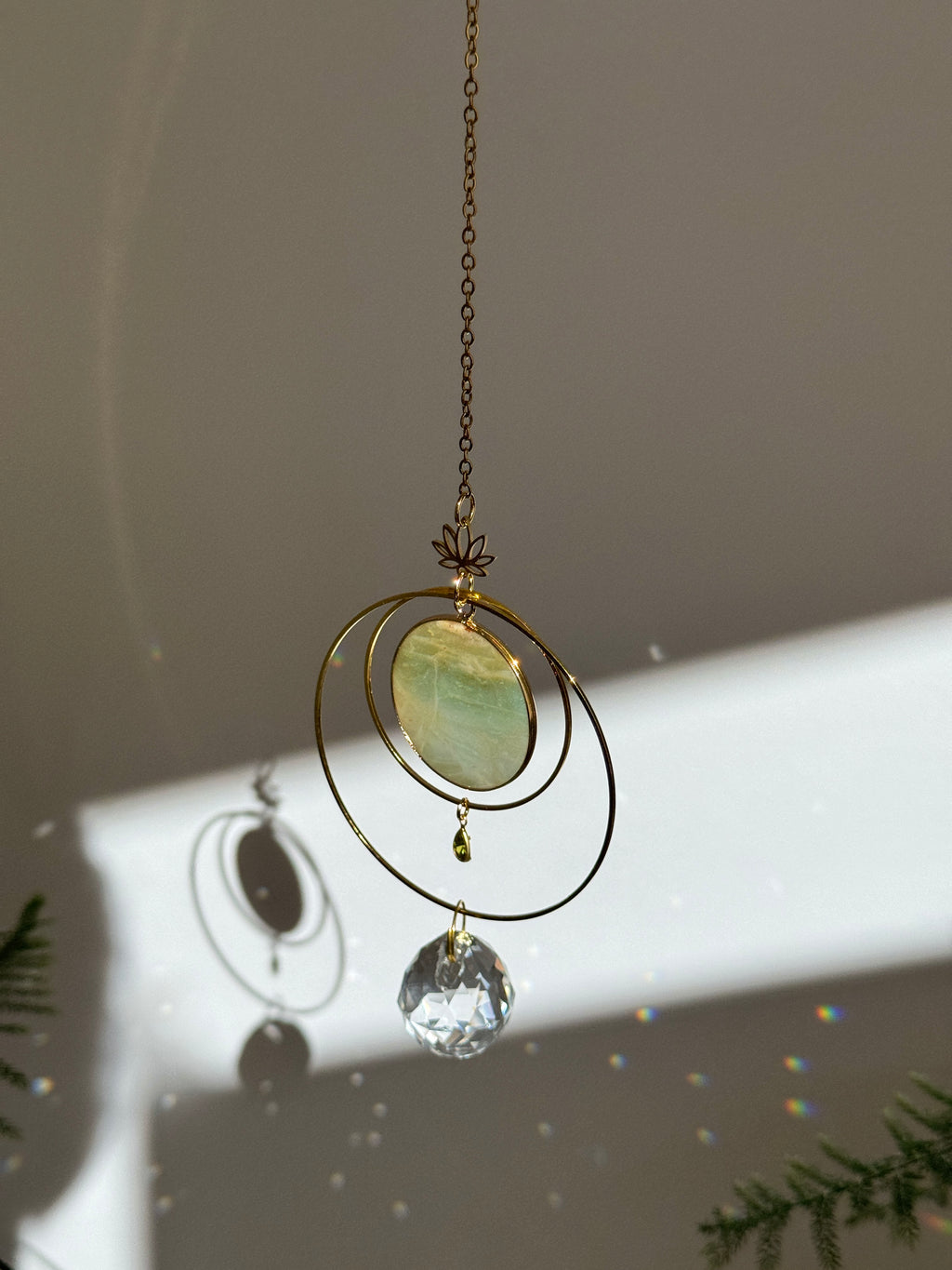Detailed view of a gold lotus suncatcher featuring a round green Amazonite stone disc and a large faceted crystal drop reflecting rainbow light spots on a soft grey background.
