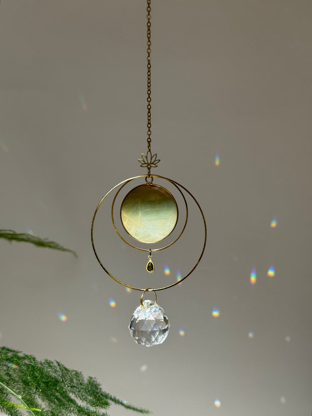 Detailed view of a gold lotus suncatcher featuring a round green Amazonite stone disc and a large faceted crystal drop reflecting rainbow light spots on a soft grey background.