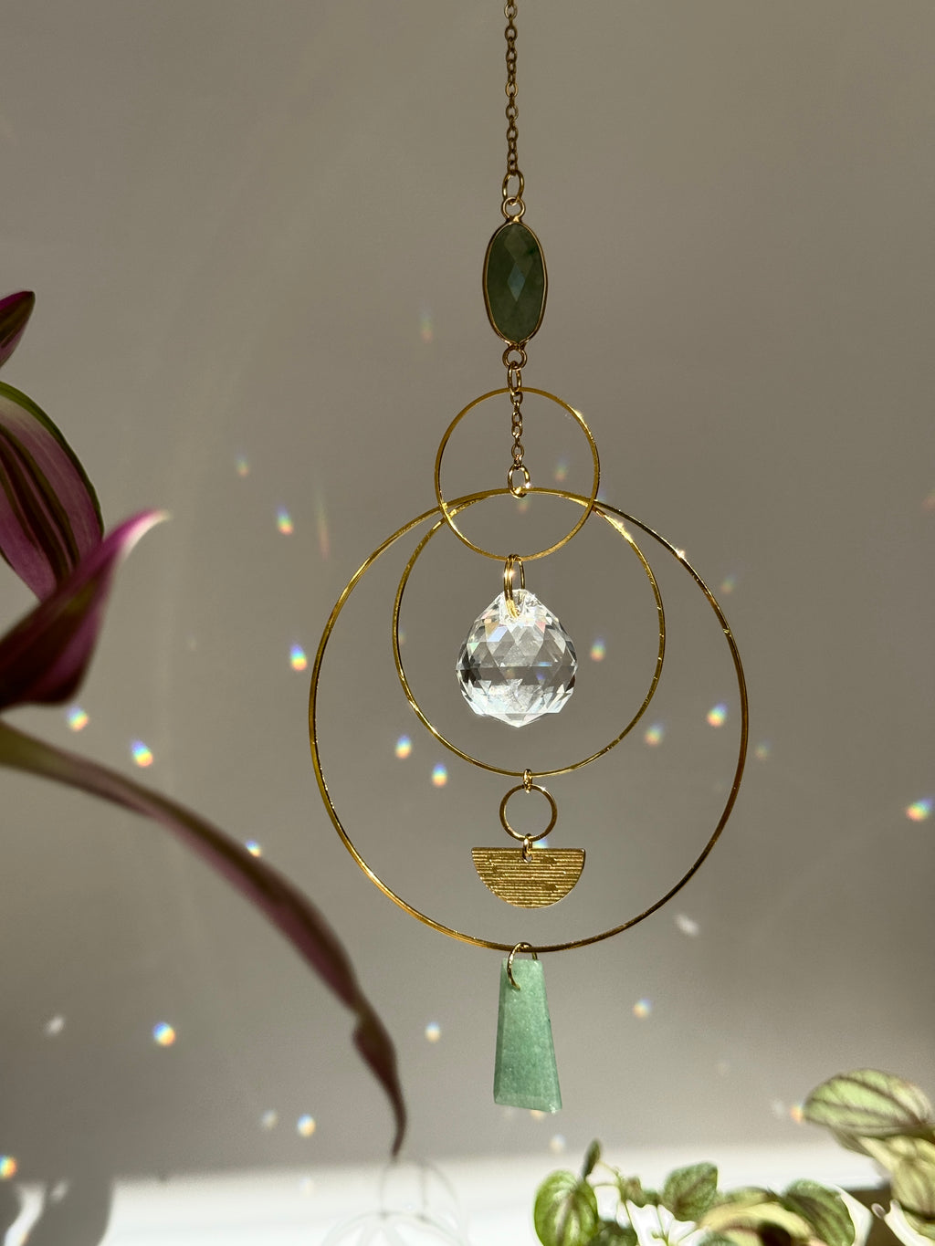 Vertical gold geometric suncatcher with three tiered hoops, a faceted crystal sphere, and two green Aventurine stones casting a galaxy of rainbows on a white wall.