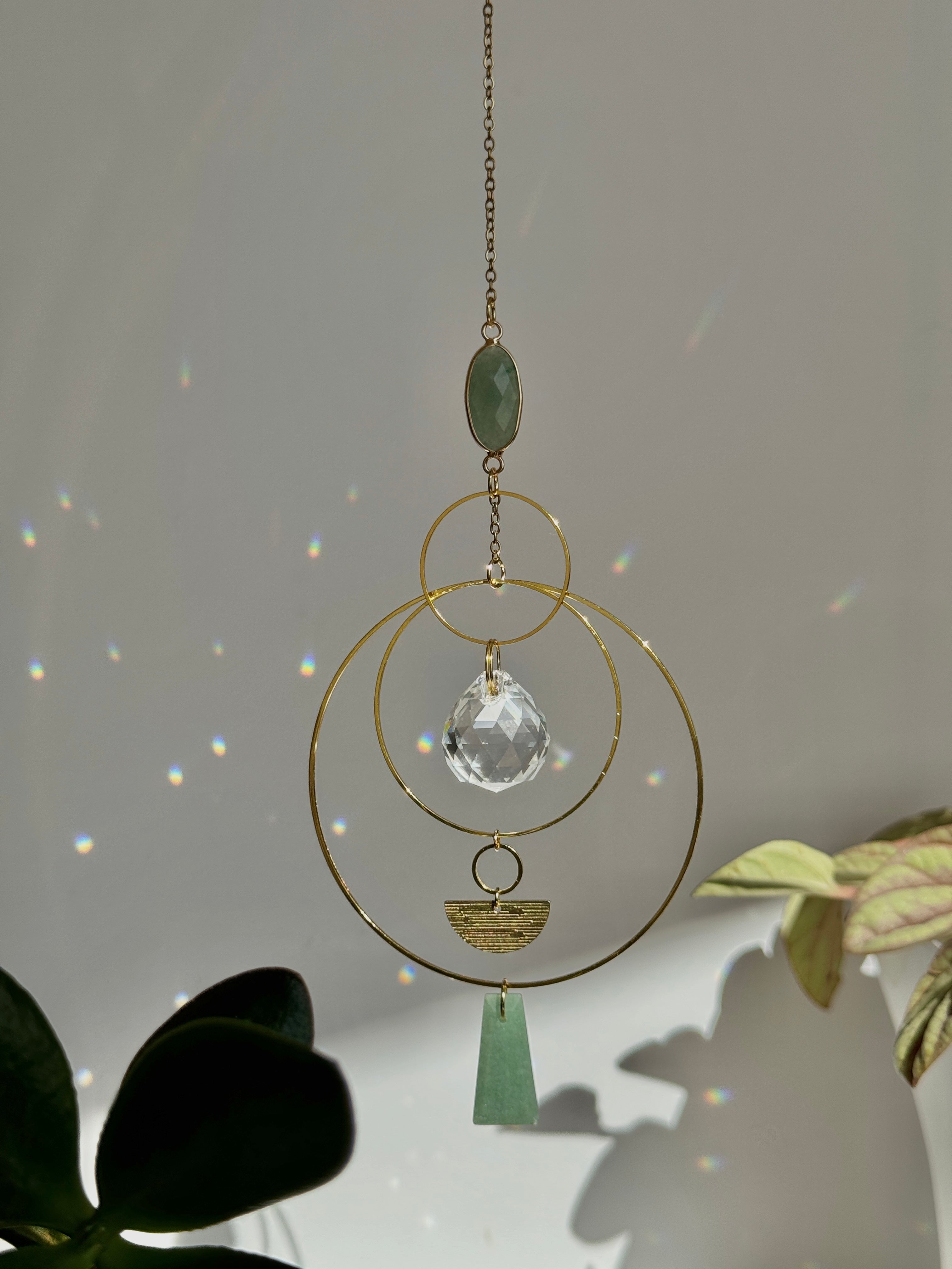 Vertical gold geometric suncatcher with three tiered hoops, a faceted crystal sphere, and two green Aventurine stones casting a galaxy of rainbows on a white wall.