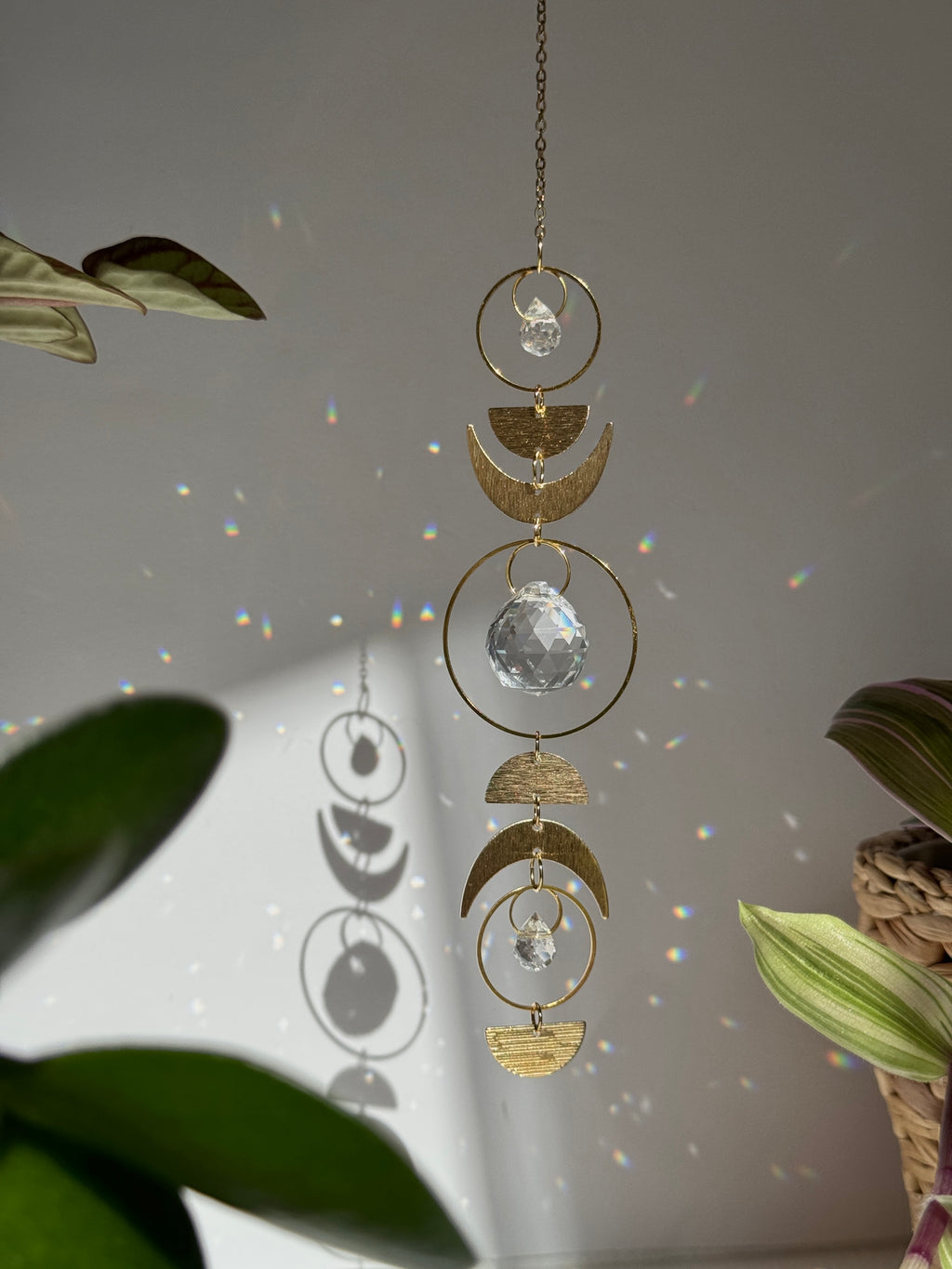 Handcrafted gold moon phase suncatcher featuring a vertical alignment of textured brass crescent moons and hoops with three clear crystal prisms casting rainbows on a white wall.
