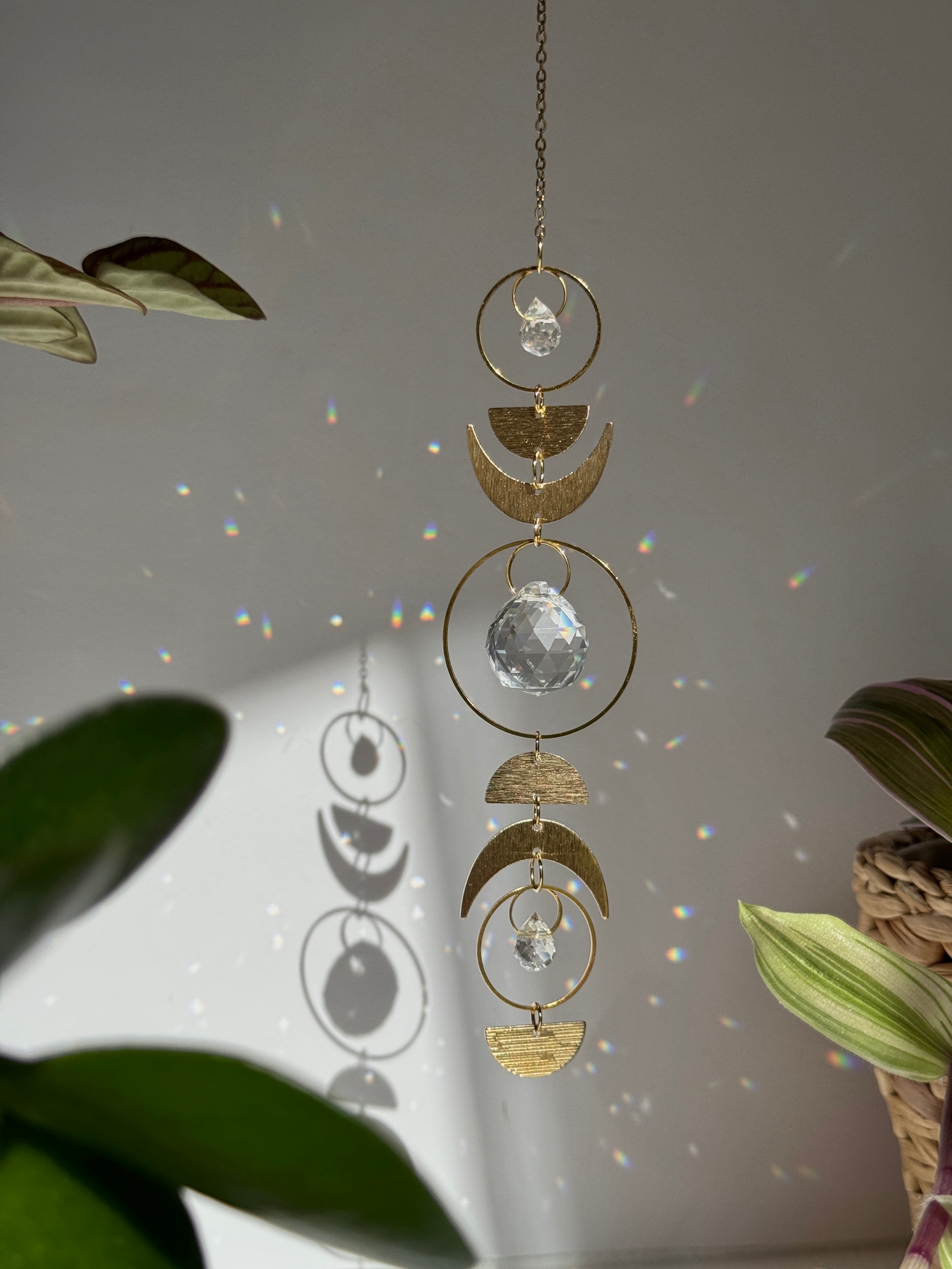 Handcrafted gold moon phase suncatcher featuring a vertical alignment of textured brass crescent moons and hoops with three clear crystal prisms casting rainbows on a white wall.