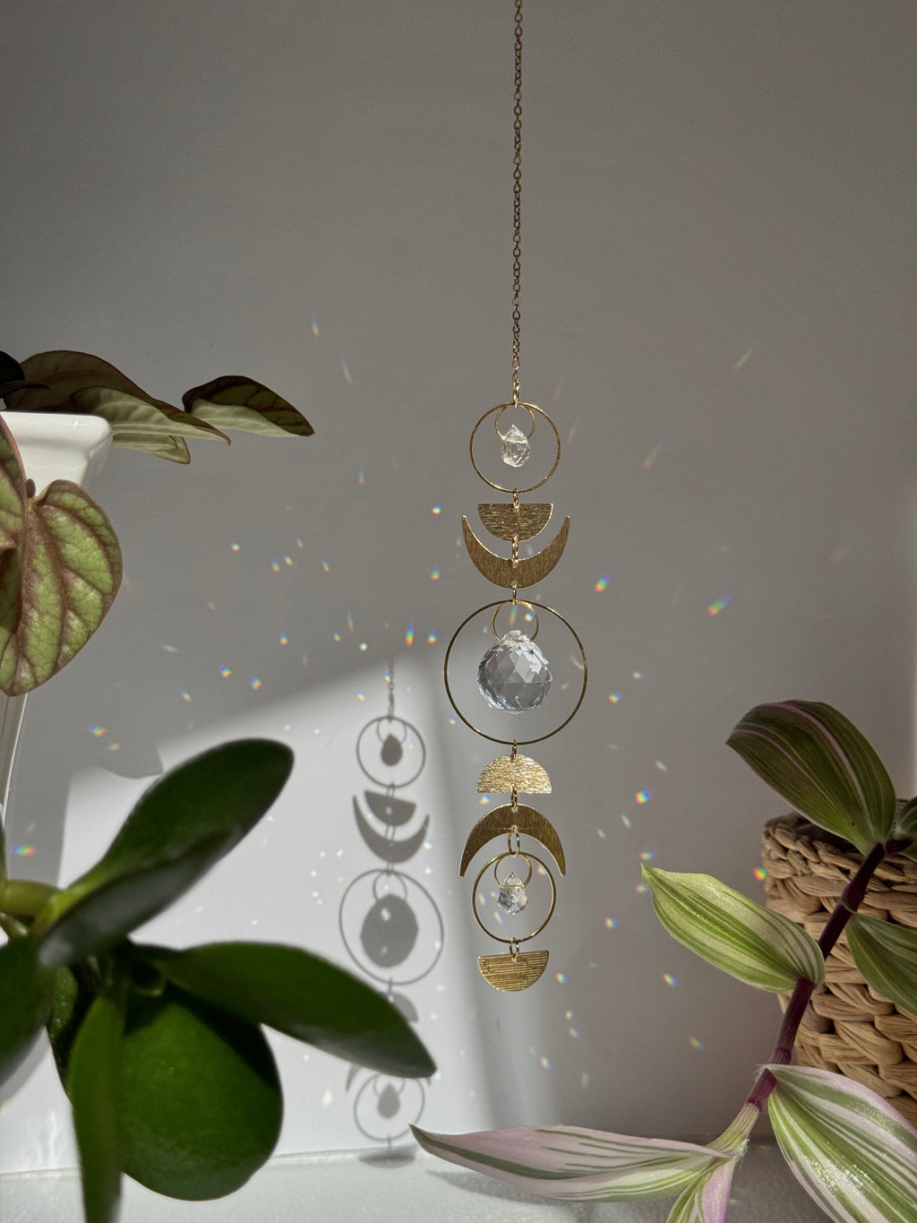 Handcrafted gold moon phase suncatcher featuring a vertical alignment of textured brass crescent moons and hoops with three clear crystal prisms casting rainbows on a white wall.