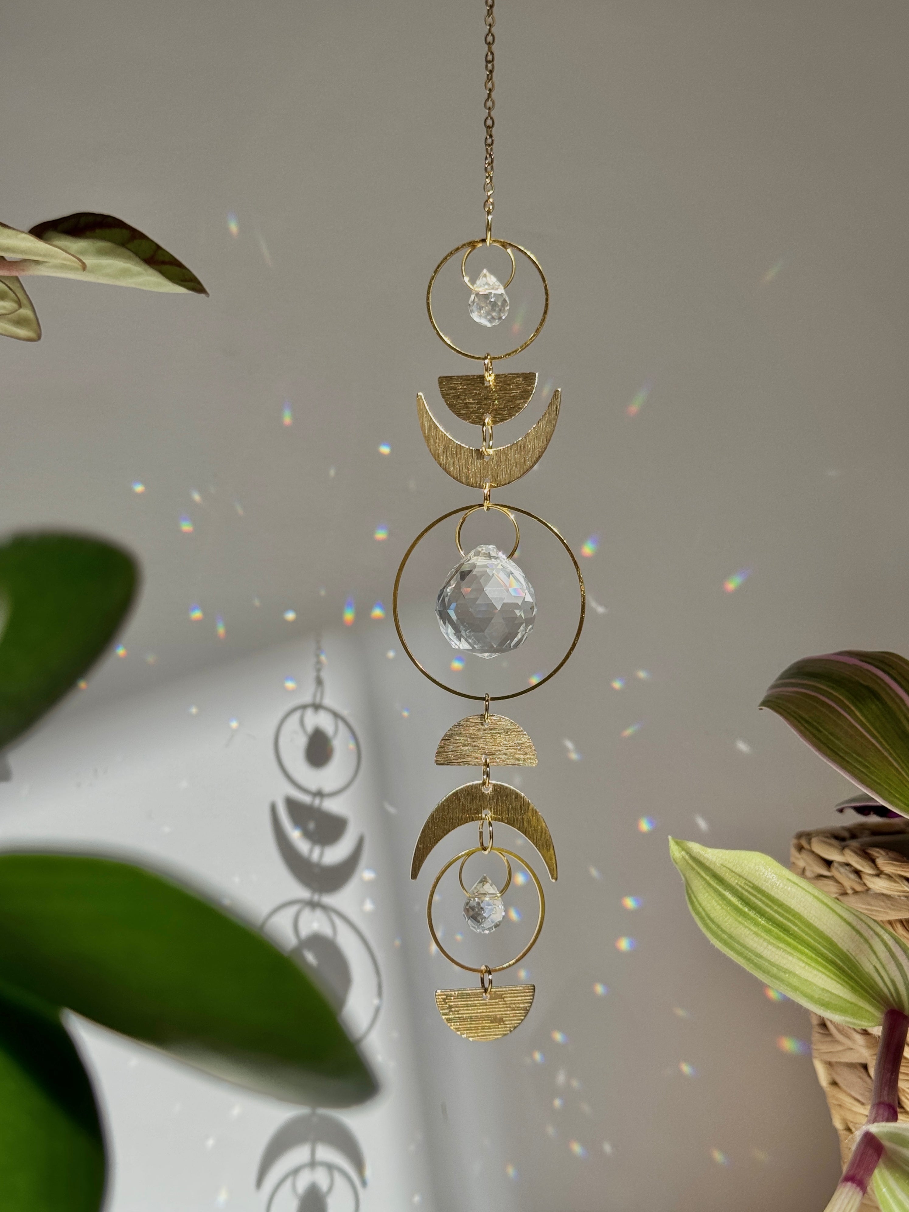 Handcrafted gold moon phase suncatcher featuring a vertical alignment of textured brass crescent moons and hoops with three clear crystal prisms casting rainbows on a white wall.