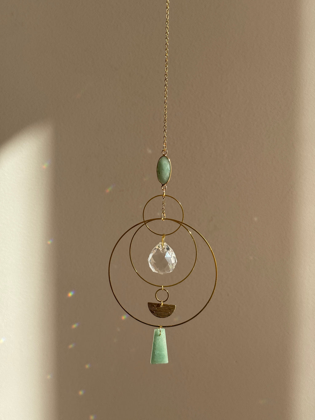 Vertical gold geometric suncatcher with three tiered hoops, a faceted crystal sphere, and two green Aventurine stones casting a galaxy of rainbows on a white wall.