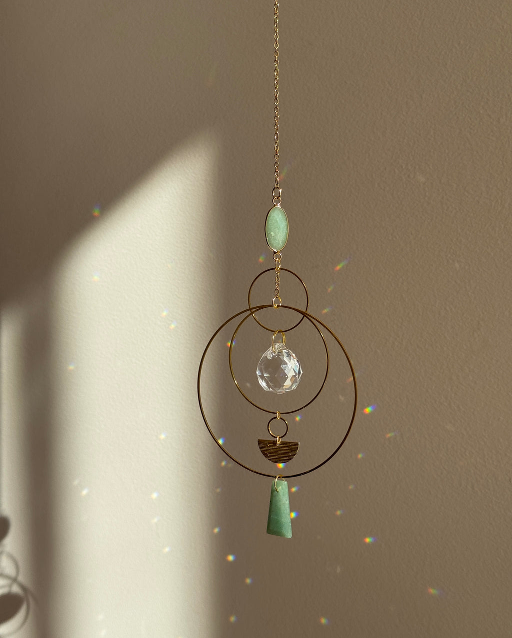 Vertical gold geometric suncatcher with three tiered hoops, a faceted crystal sphere, and two green Aventurine stones casting a galaxy of rainbows on a white wall.