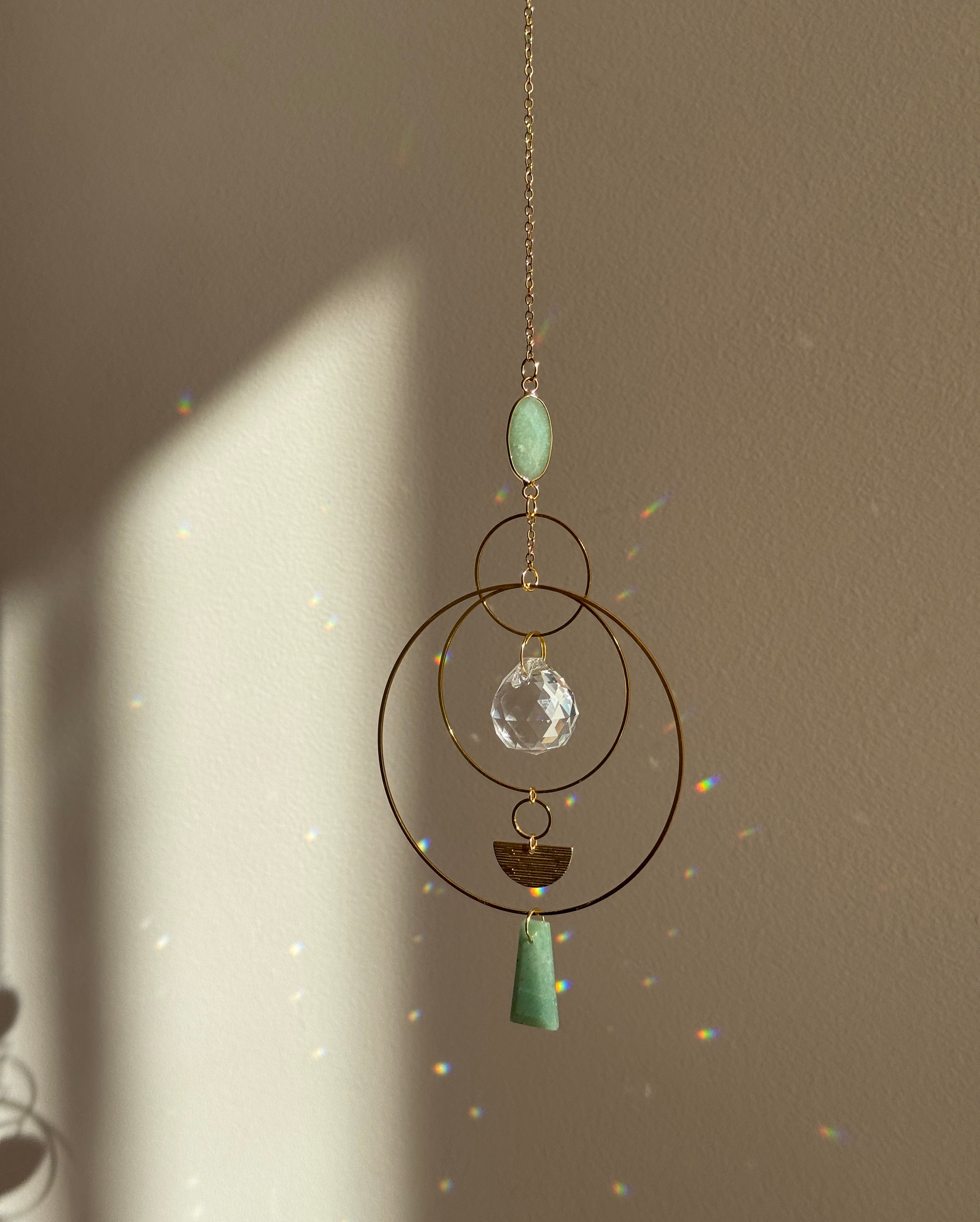 Vertical gold geometric suncatcher with three tiered hoops, a faceted crystal sphere, and two green Aventurine stones casting a galaxy of rainbows on a white wall.