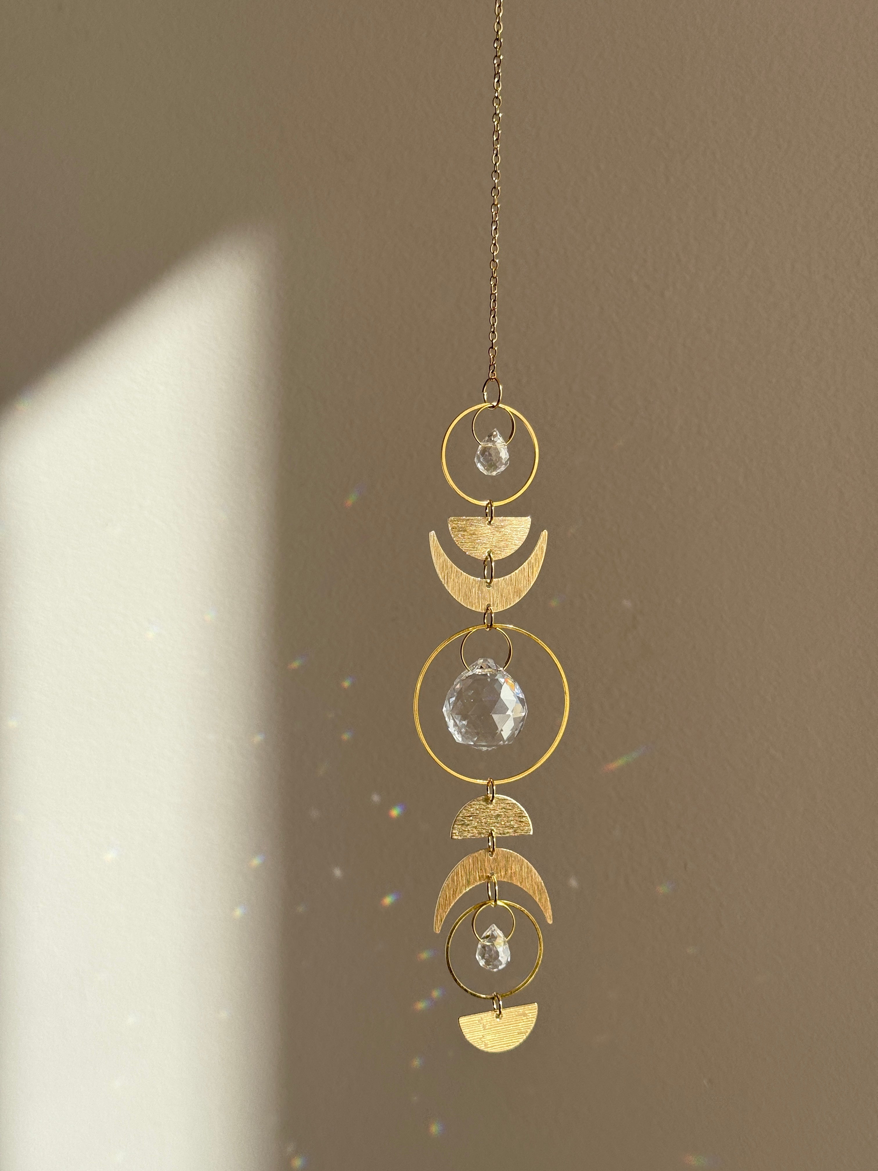 Handcrafted gold moon phase suncatcher featuring a vertical alignment of textured brass crescent moons and hoops with three clear crystal prisms casting rainbows on a white wall.