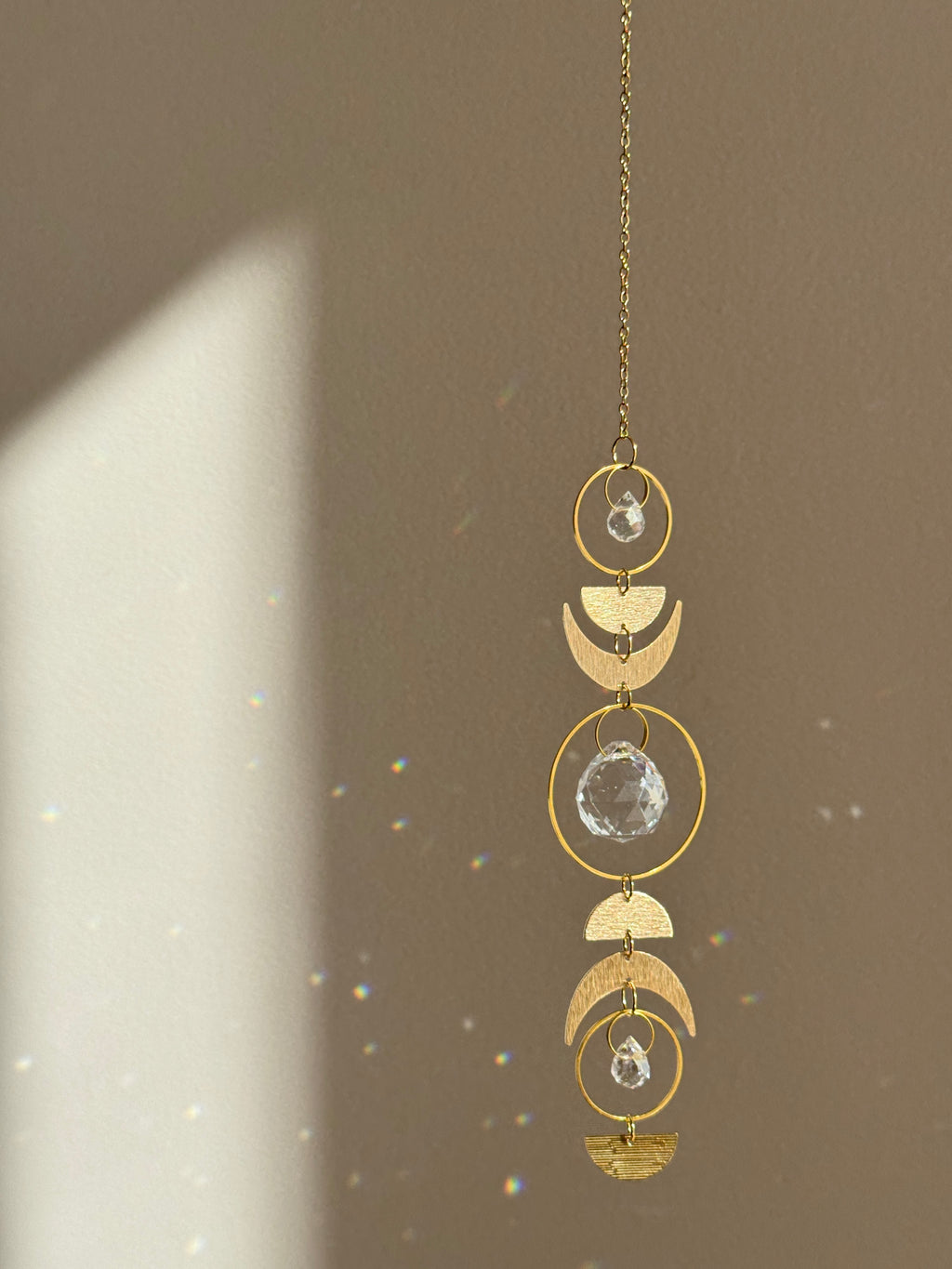 Handcrafted gold moon phase suncatcher featuring a vertical alignment of textured brass crescent moons and hoops with three clear crystal prisms casting rainbows on a white wall.