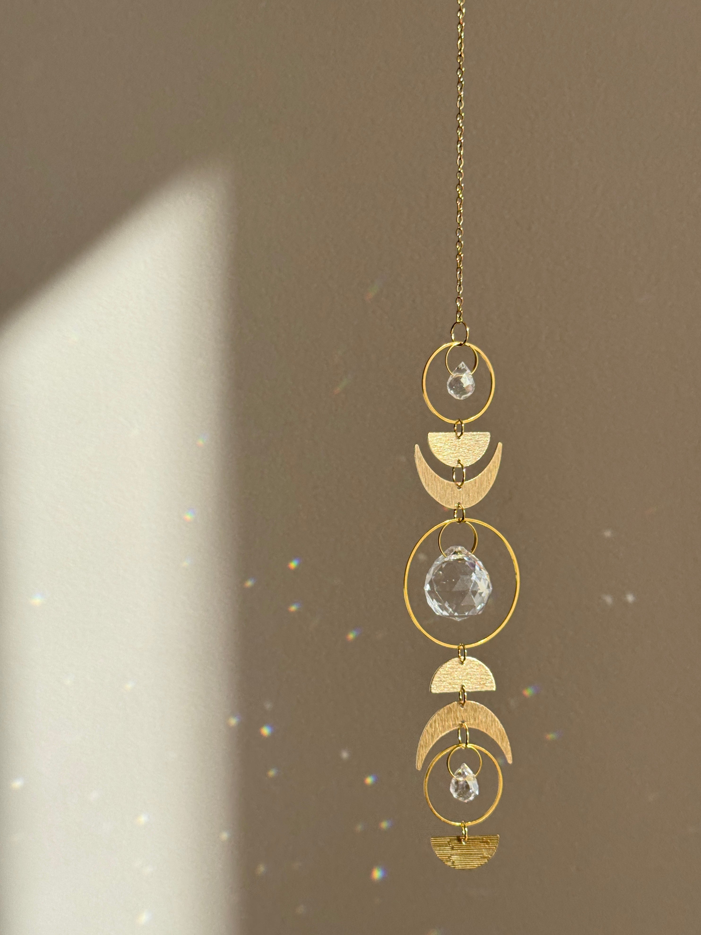 Handcrafted gold moon phase suncatcher featuring a vertical alignment of textured brass crescent moons and hoops with three clear crystal prisms casting rainbows on a white wall.