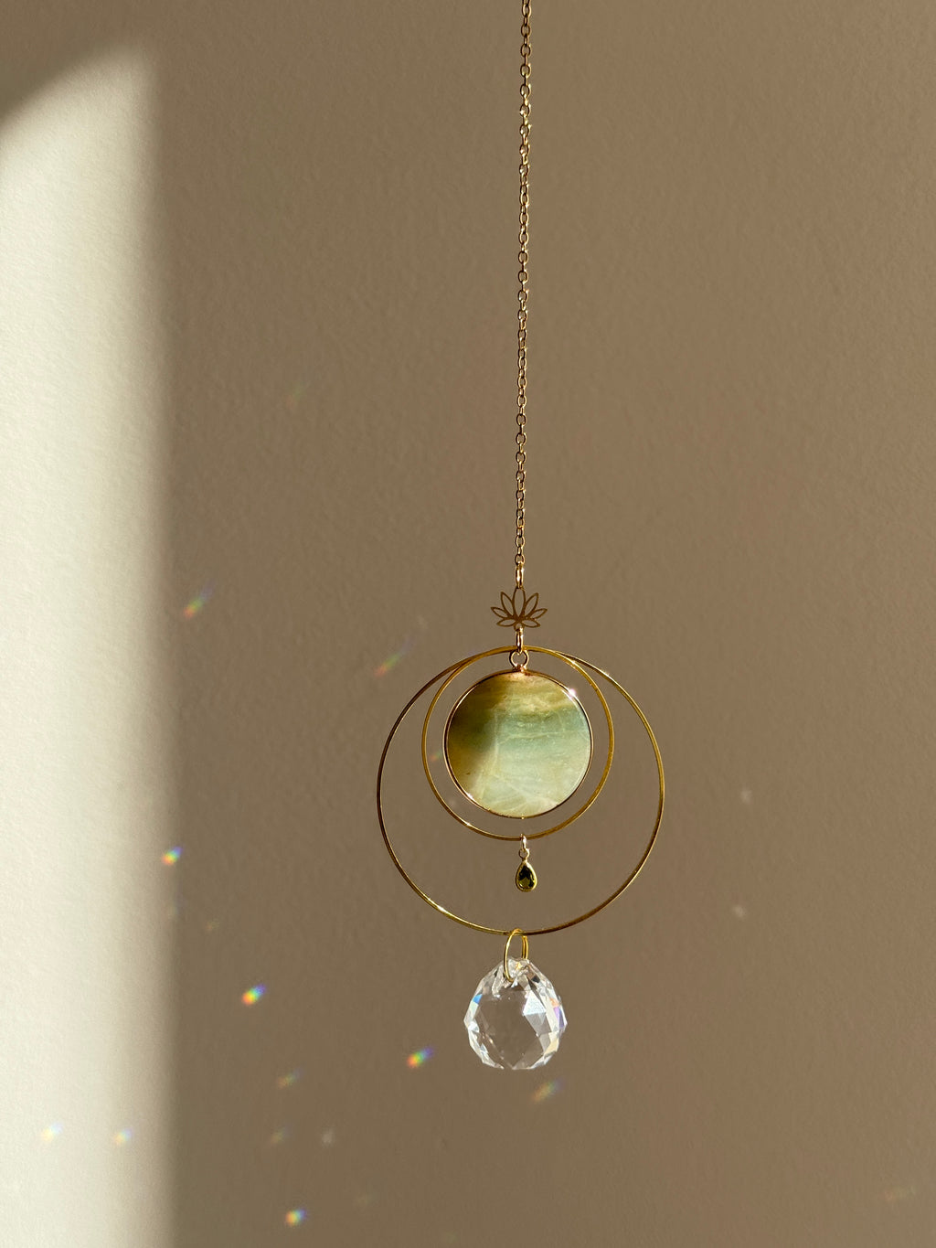 Detailed view of a gold lotus suncatcher featuring a round green Amazonite stone disc and a large faceted crystal drop reflecting rainbow light spots on a soft grey background.
