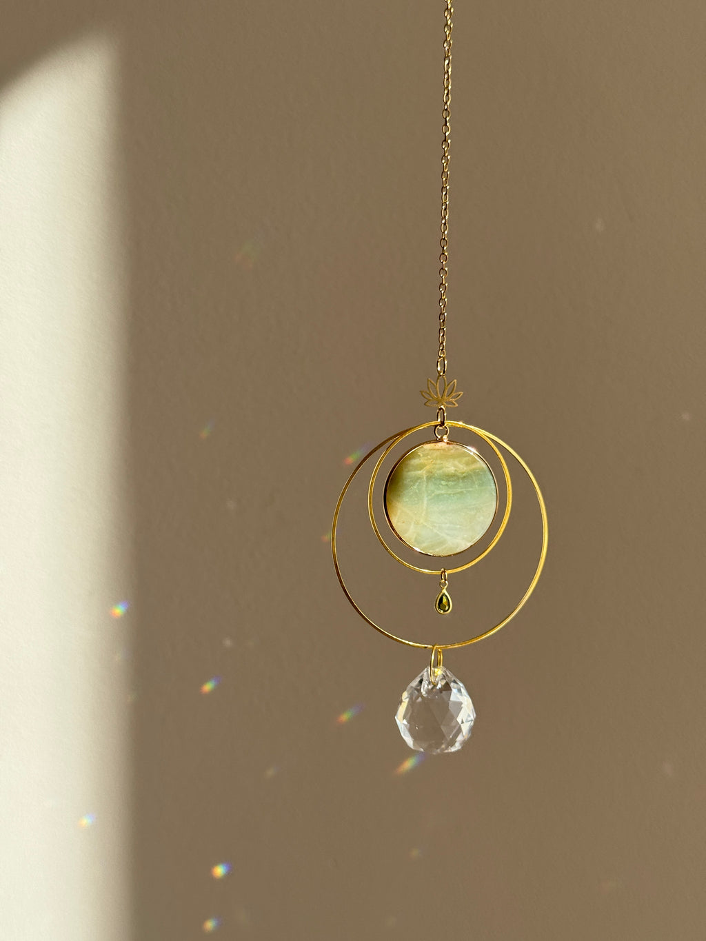 Detailed view of a gold lotus suncatcher featuring a round green Amazonite stone disc and a large faceted crystal drop reflecting rainbow light spots on a soft grey background.