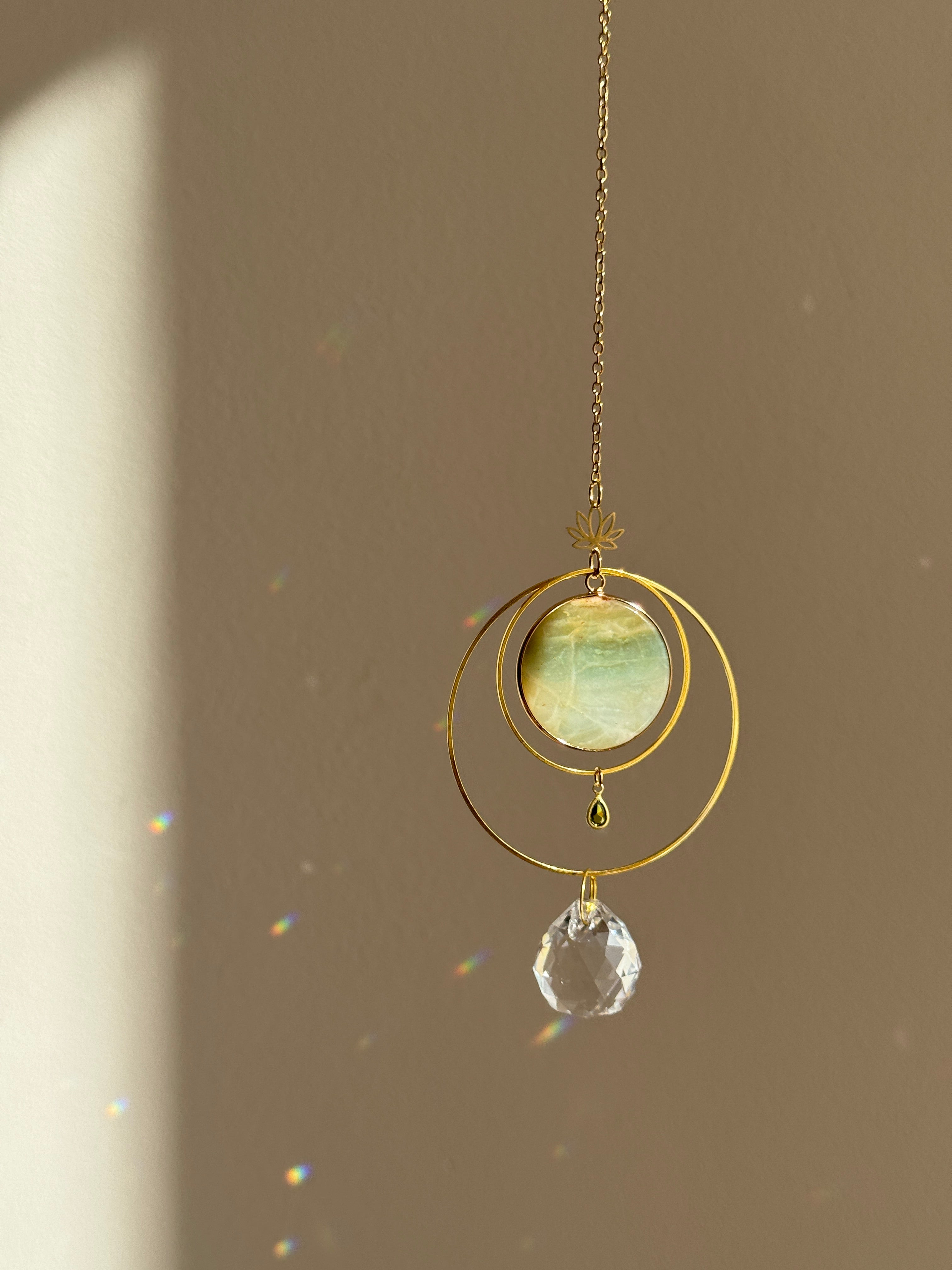 Detailed view of a gold lotus suncatcher featuring a round green Amazonite stone disc and a large faceted crystal drop reflecting rainbow light spots on a soft grey background.