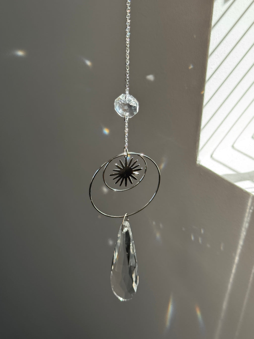 Handmade silver suncatcher with a minimalist starburst design and a large faceted teardrop crystal prism, hanging in natural sunlight with rainbow reflections and shadows on a grey wall background.