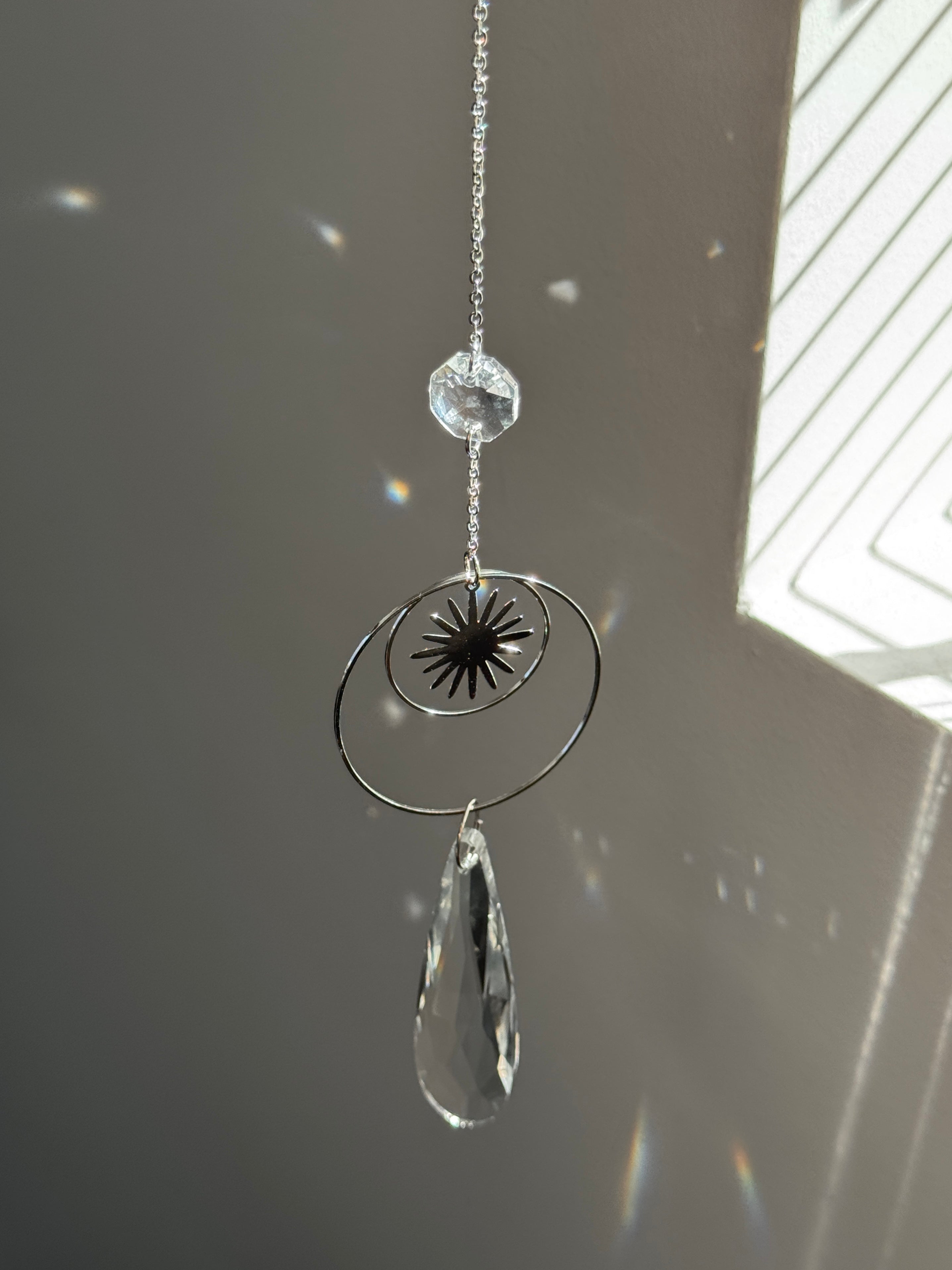 Handmade silver suncatcher with a minimalist starburst design and a large faceted teardrop crystal prism, hanging in natural sunlight with rainbow reflections and shadows on a grey wall background.