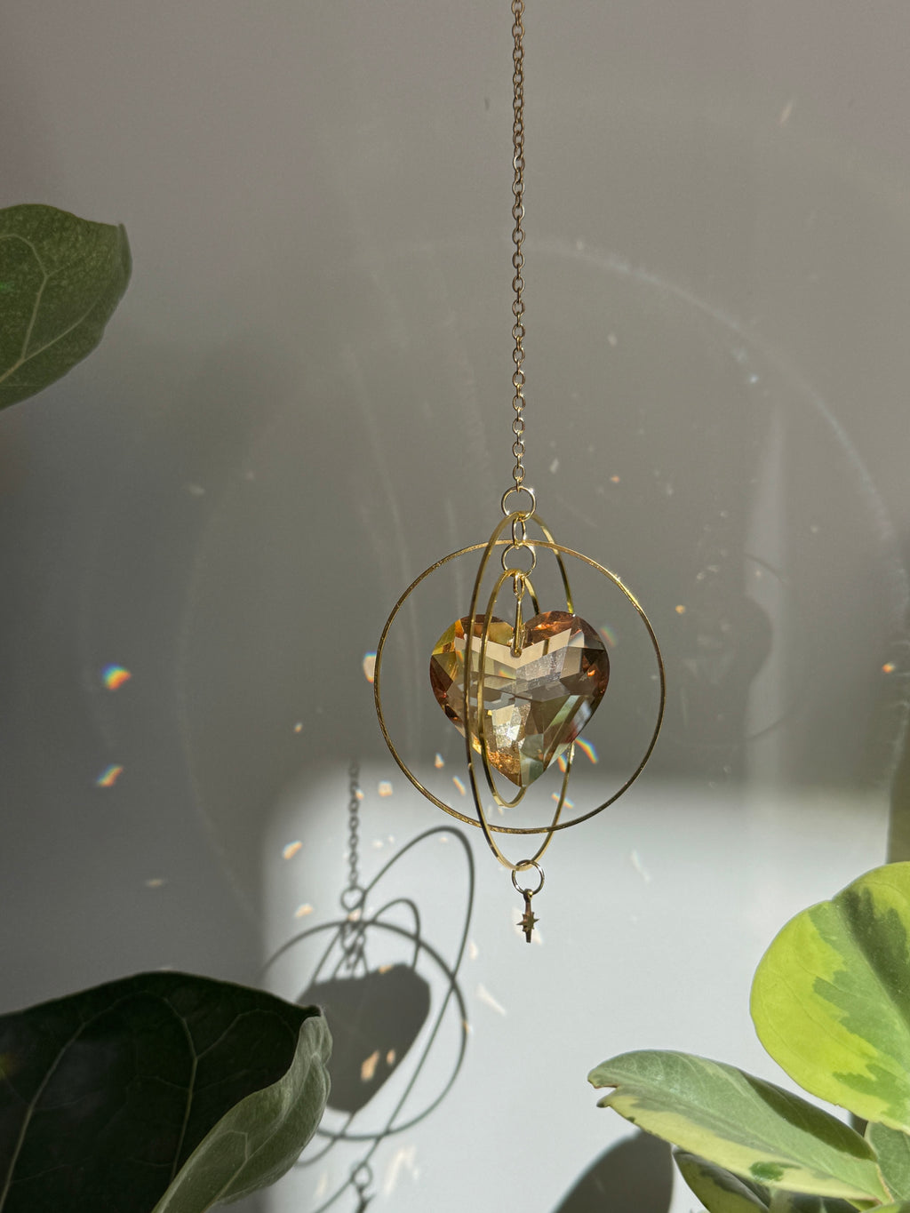 Sunkissed Heartbeat crystal suncatcher featuring a warm amber-gold faceted heart prism. Handmade celestial window decor with gold rings casting rainbow light flares (Sonnenfänger).