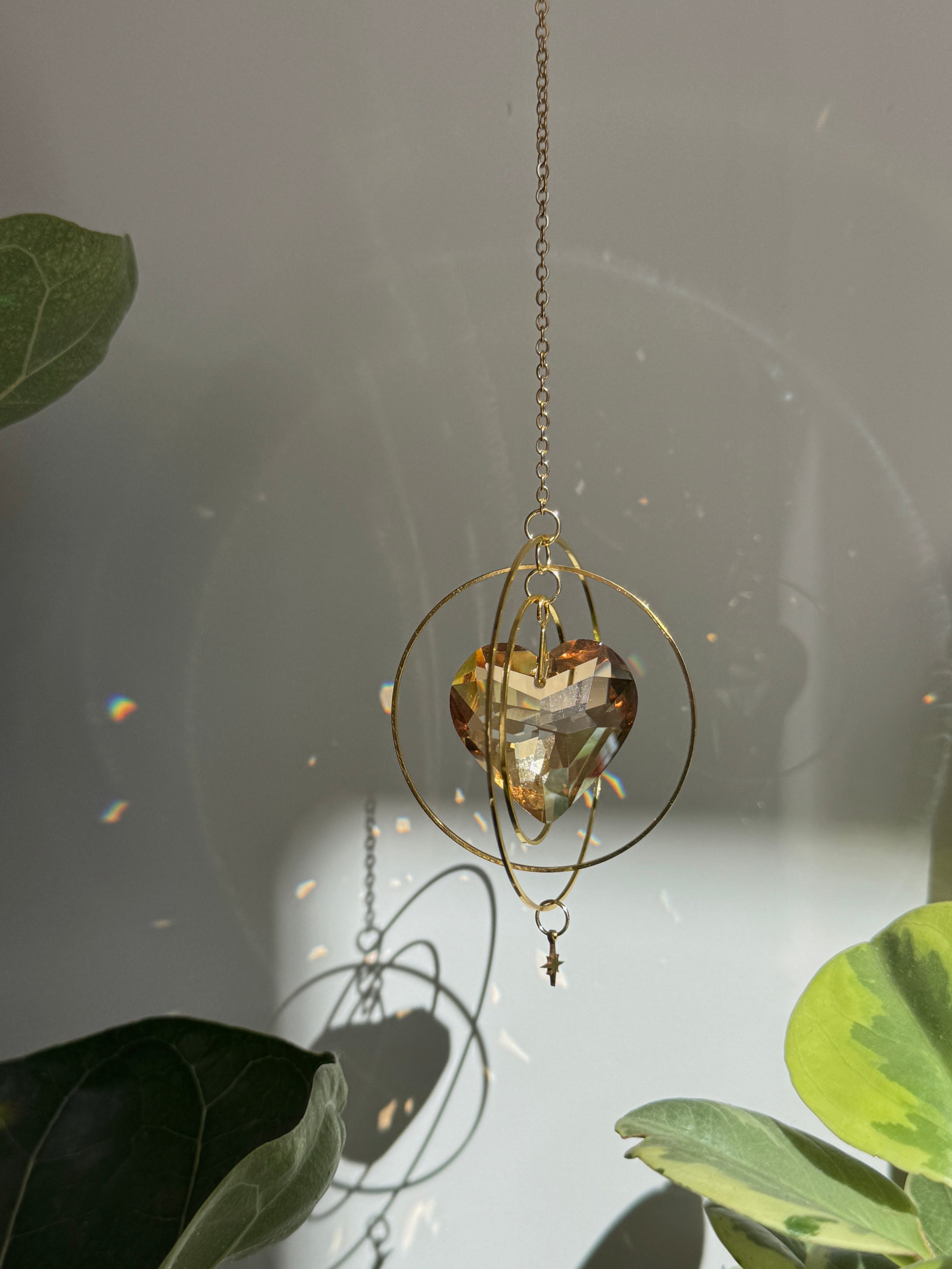 Sunkissed Heartbeat crystal suncatcher featuring a warm amber-gold faceted heart prism. Handmade celestial window decor with gold rings casting rainbow light flares (Sonnenfänger).