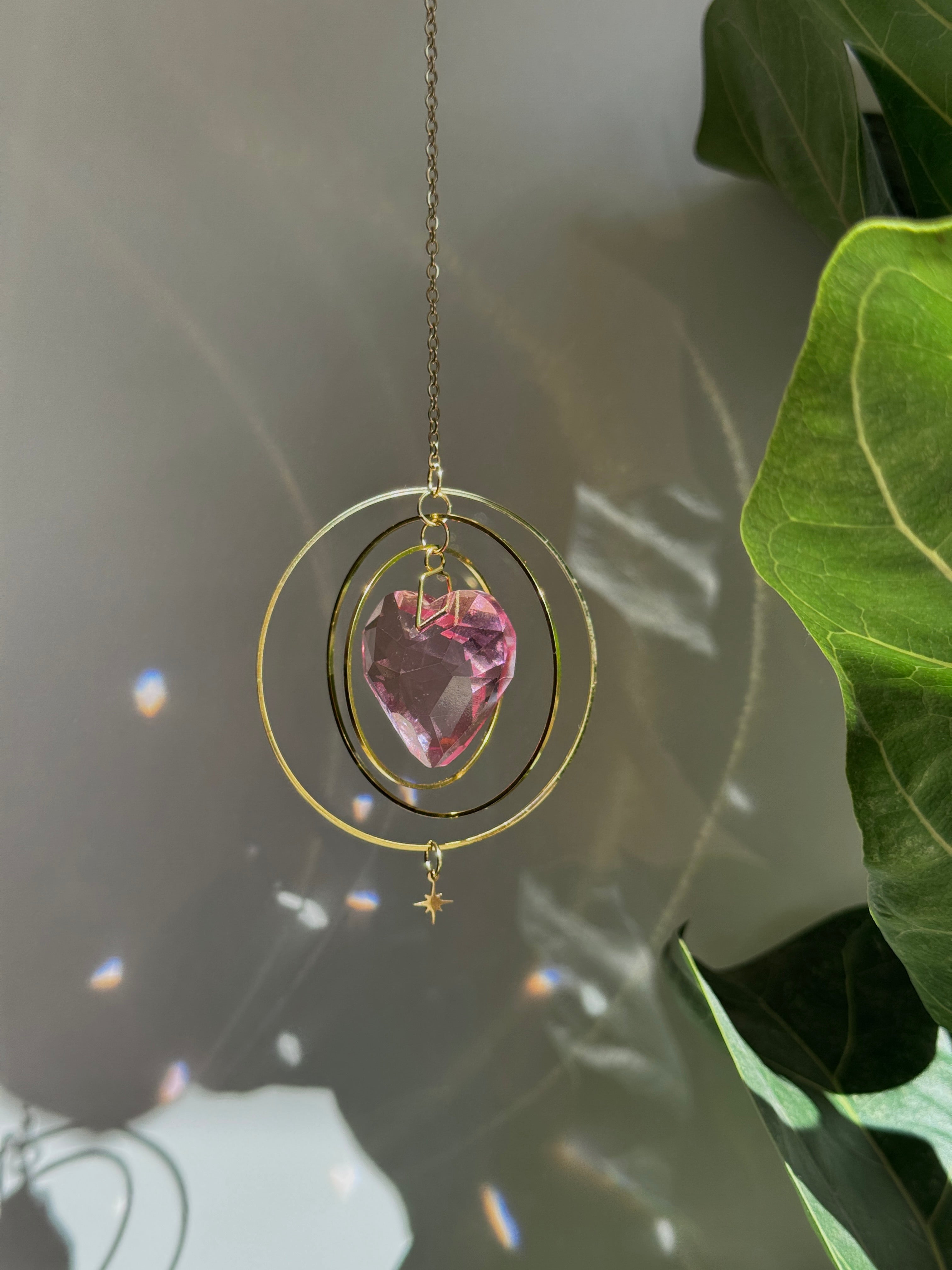 Sunkissed Heartbeat crystal suncatcher featuring a pink-gold faceted heart prism. Handmade celestial window decor with gold rings casting rainbow light flares (Sonnenfänger).