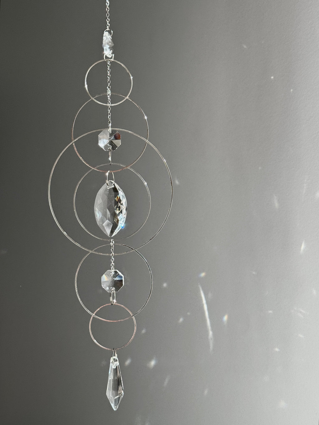 60cm silver geometric suncatcher featuring interlocking concentric hoops and clear marquise-cut crystal prisms, hanging in a window and casting rainbows on a wall.