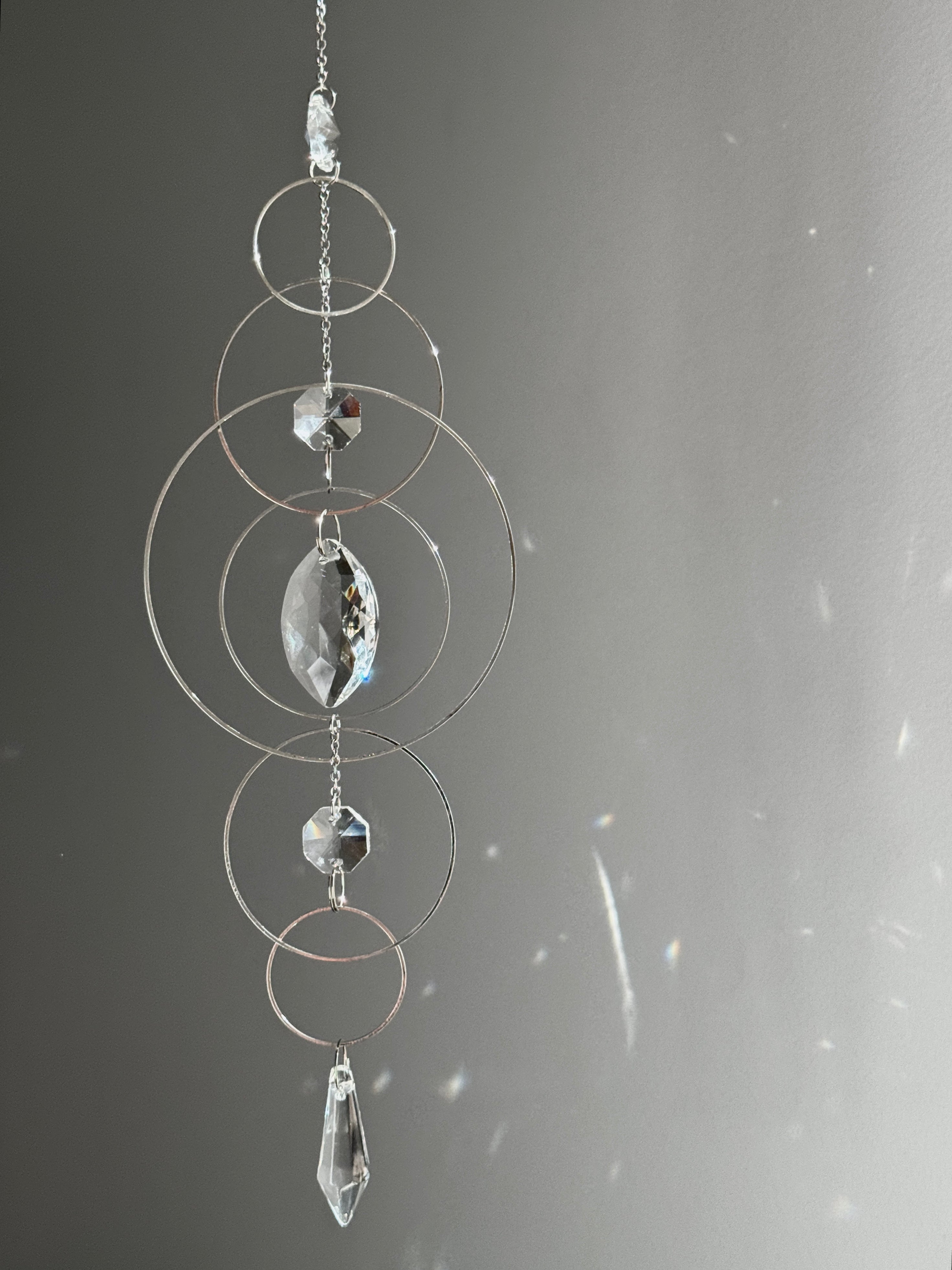 60cm silver geometric suncatcher featuring interlocking concentric hoops and clear marquise-cut crystal prisms, hanging in a window and casting rainbows on a wall.