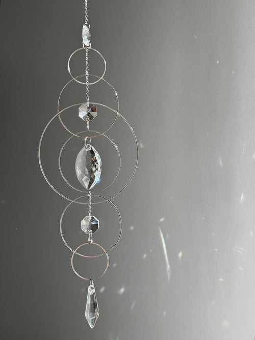 60cm silver geometric suncatcher featuring interlocking concentric hoops and clear marquise-cut crystal prisms, hanging in a window and casting rainbows on a wall.