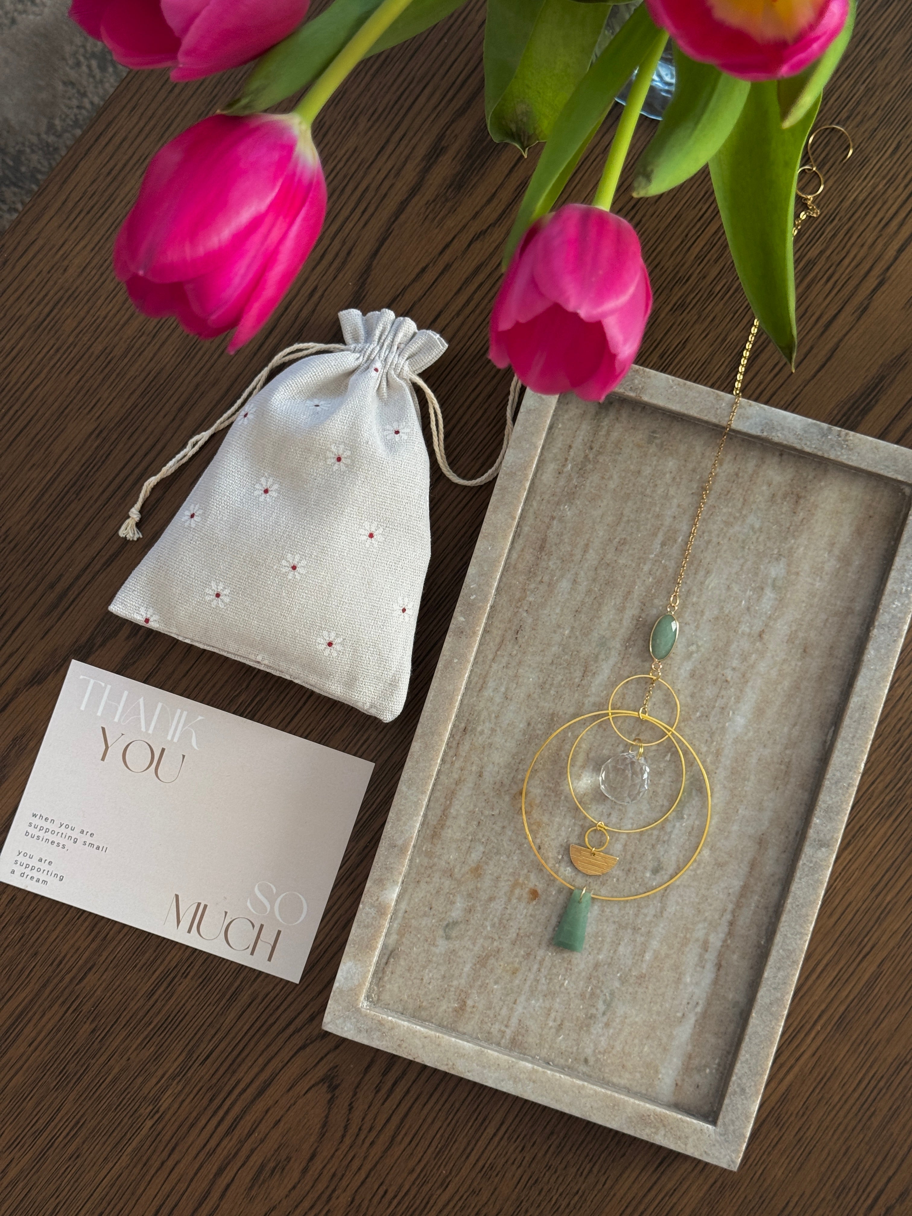 Vertical gold geometric suncatcher with three tiered hoops, a faceted crystal sphere, and two green Aventurine stones casting a galaxy of rainbows on a white wall.