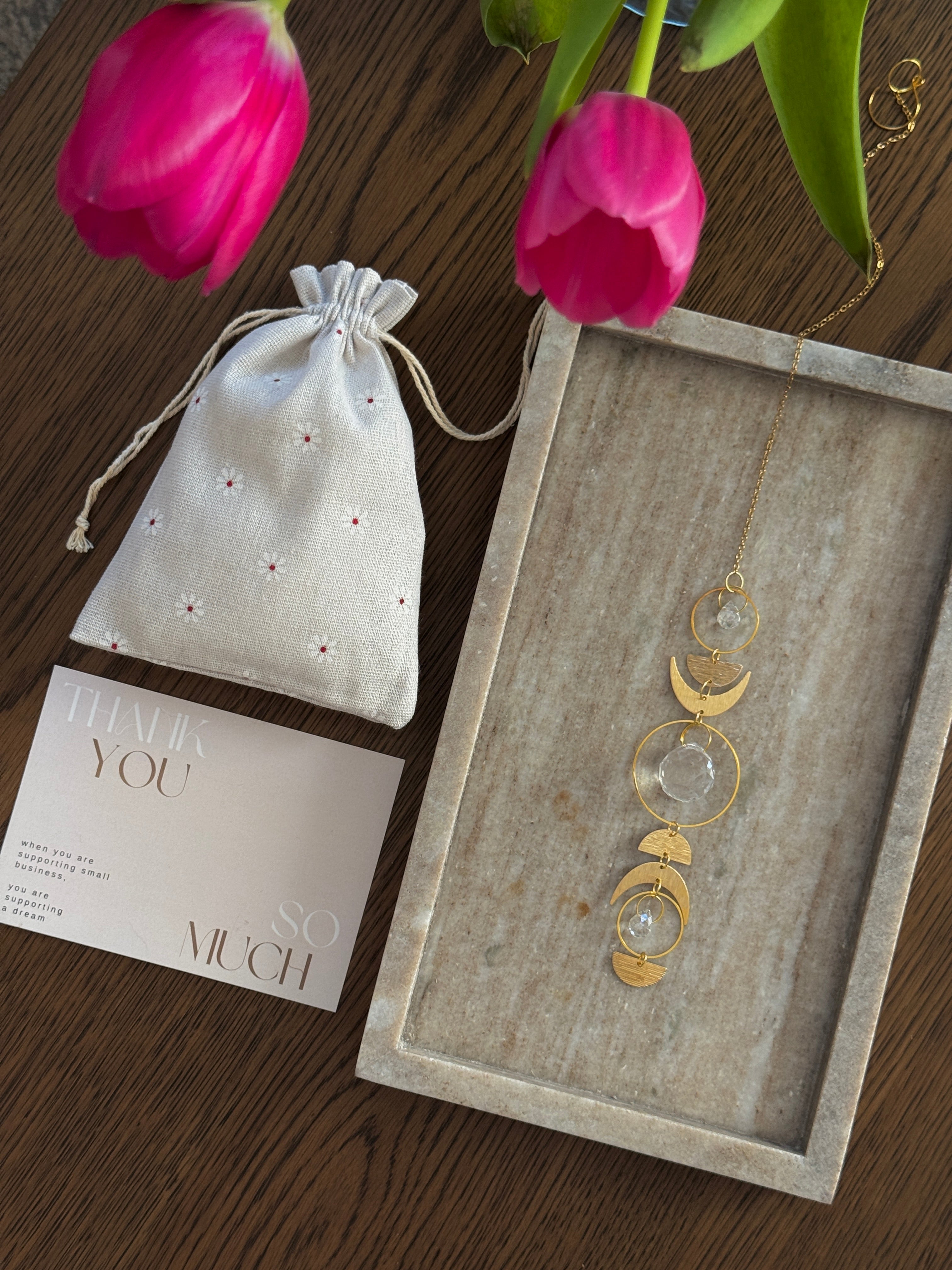 Handcrafted gold moon phase suncatcher featuring a vertical alignment of textured brass crescent moons and hoops with three clear crystal prisms casting rainbows on a white wall.