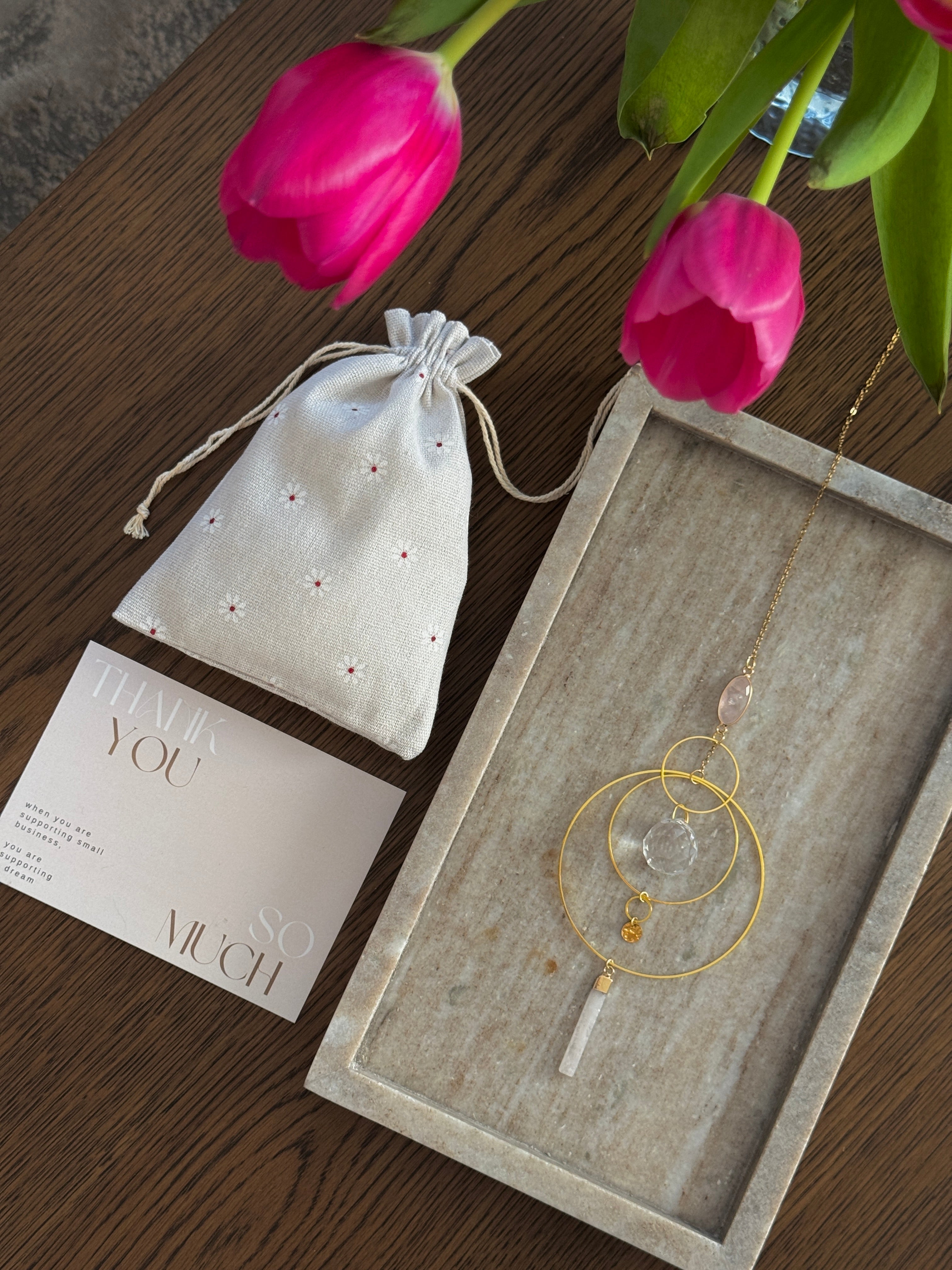 A gold geometric suncatcher featuring two pink Rose Quartz gemstones and a faceted crystal sphere. The suncatcher consists of two interlocking gold hoops, a textured coin charm, and is hanging against a soft-focus background, reflecting bright light.