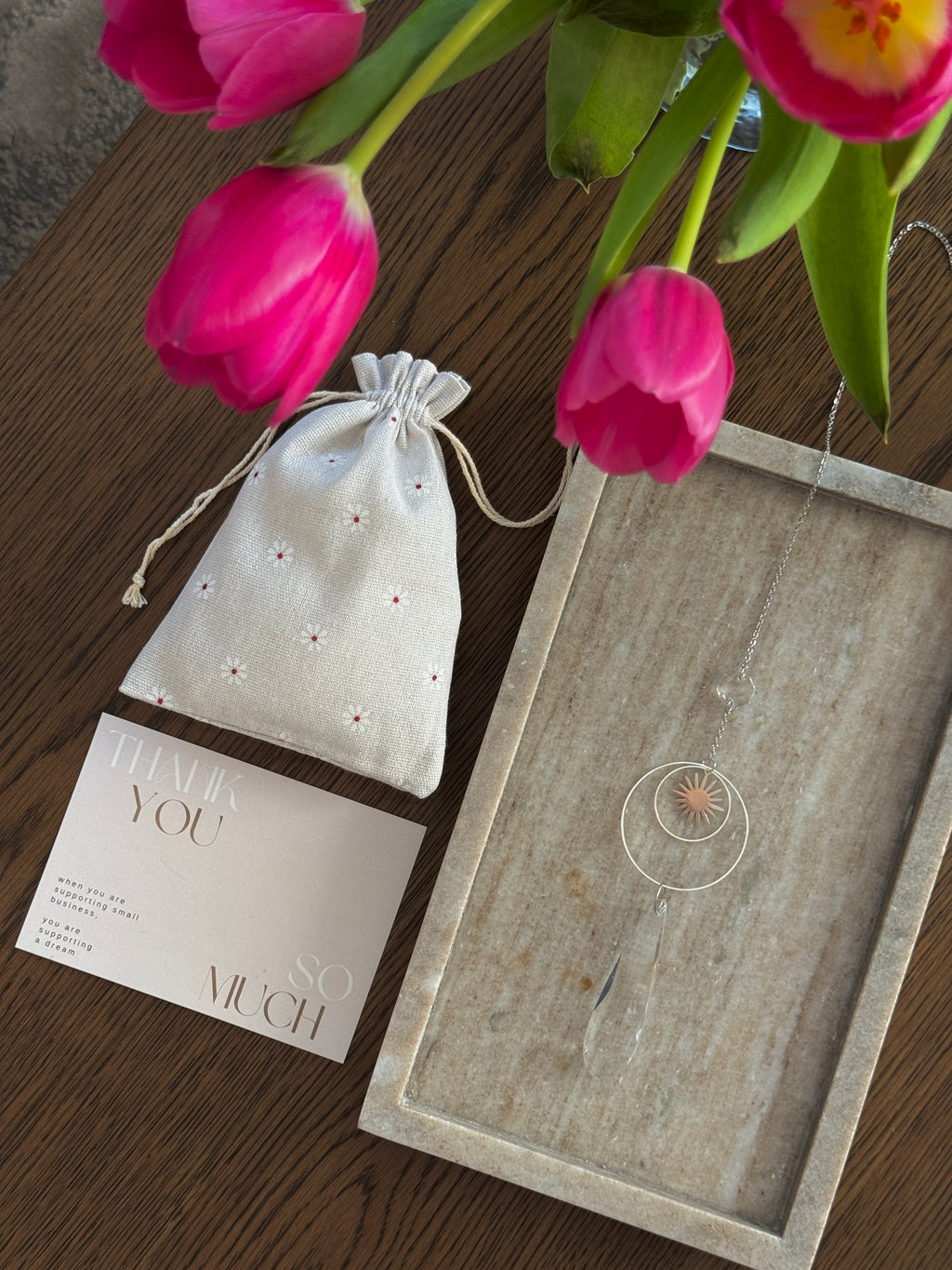 Handmade silver suncatcher with a minimalist starburst design and a large faceted teardrop crystal prism, hanging in natural sunlight with rainbow reflections and shadows on a grey wall background.