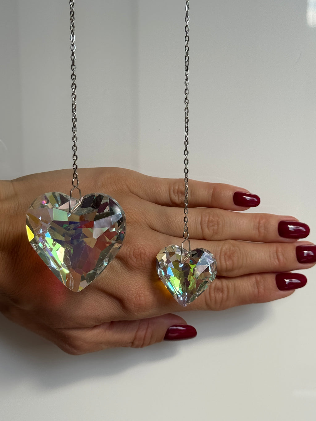 Two iridescent heart crystals, 3cm and 5cm, reflecting sunlight into colorful spectral patterns. Part of the 'Sweet Reflection' collection, showing the stainless steel hanging hook and delicate chain.